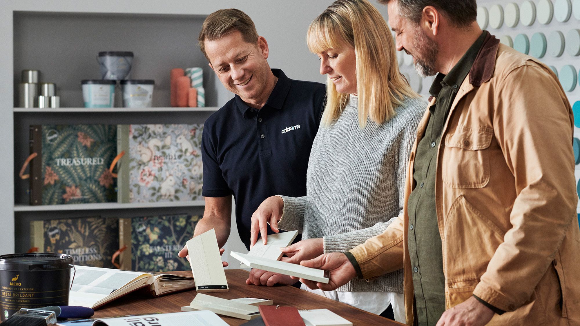 An assistant helps two customers review paint and wallpaper samples in a showroom.