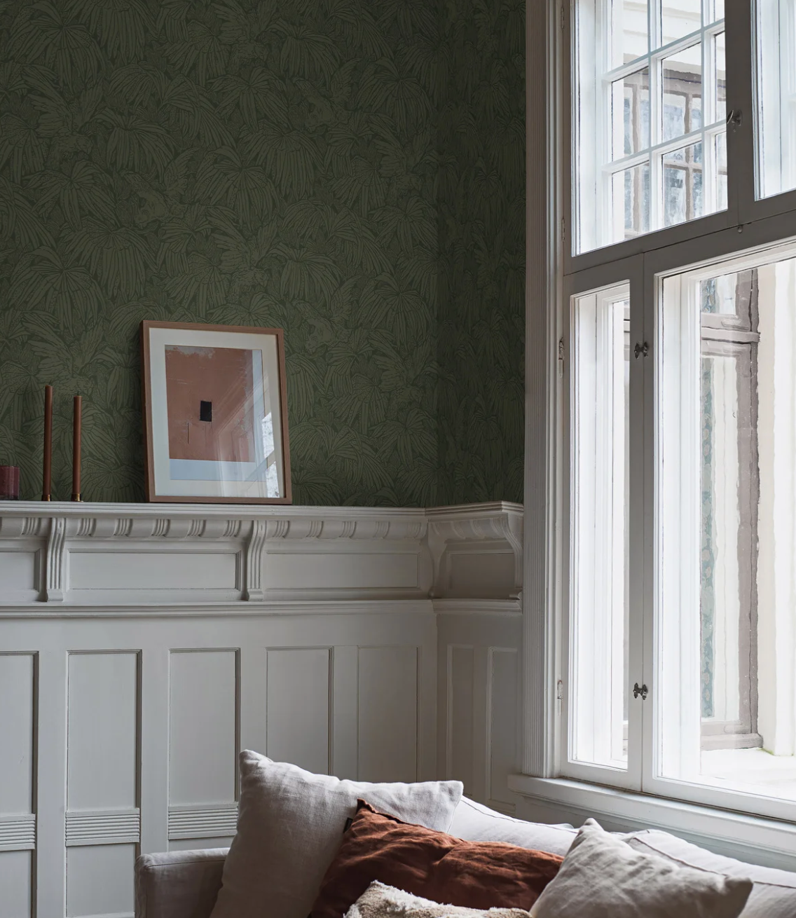 A room with dark green leaf-patterned wallpaper above white ornate wainscoting, featuring an abstract artwork on a mantel and a large multi-pane window, with a sofa in the foreground.