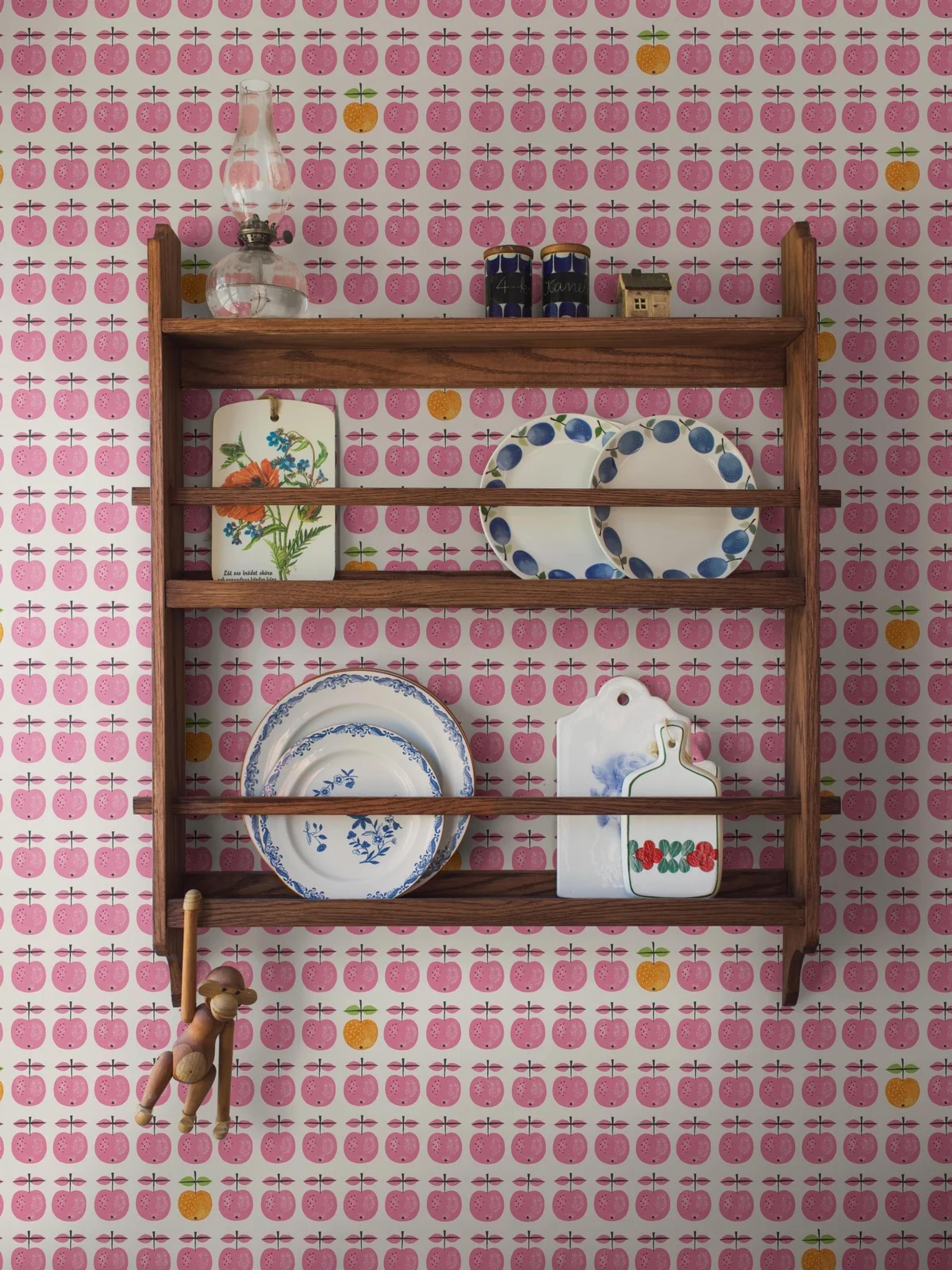 A wooden plate rack on white wallpaper patterned with pink and yellow apples, holding an oil lamp, decorative plates and plaques, and a hanging wooden monkey.