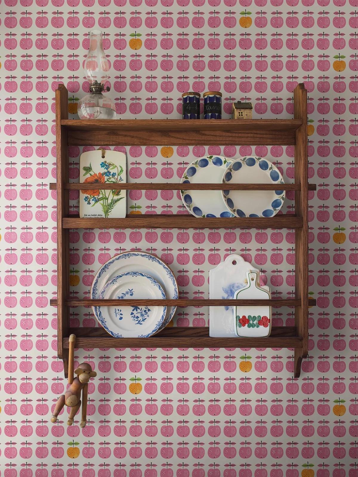 A wooden plate rack on white wallpaper patterned with pink and yellow apples, holding an oil lamp, decorative plates and plaques, and a hanging wooden monkey.