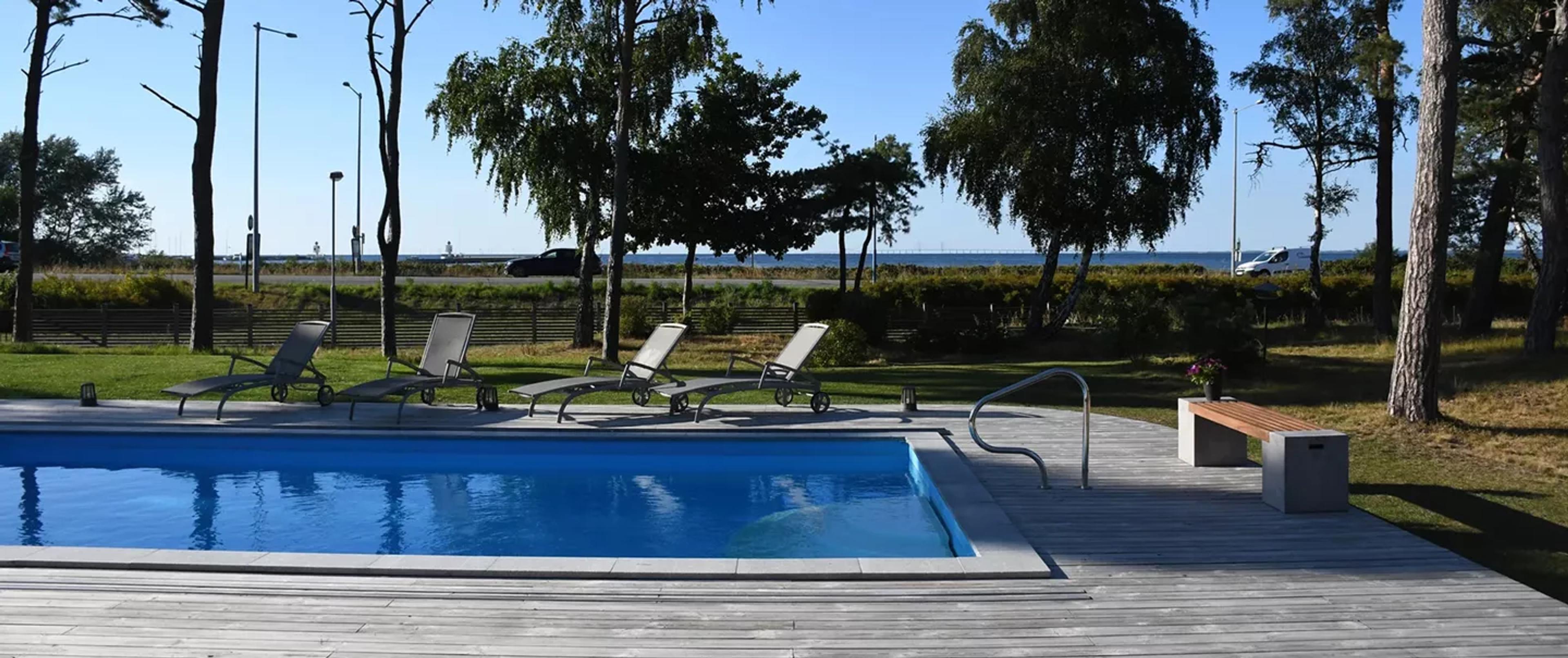 Outdoor swimming pool with lounge chairs on a wooden deck, overlooking a coastal road and the sea.