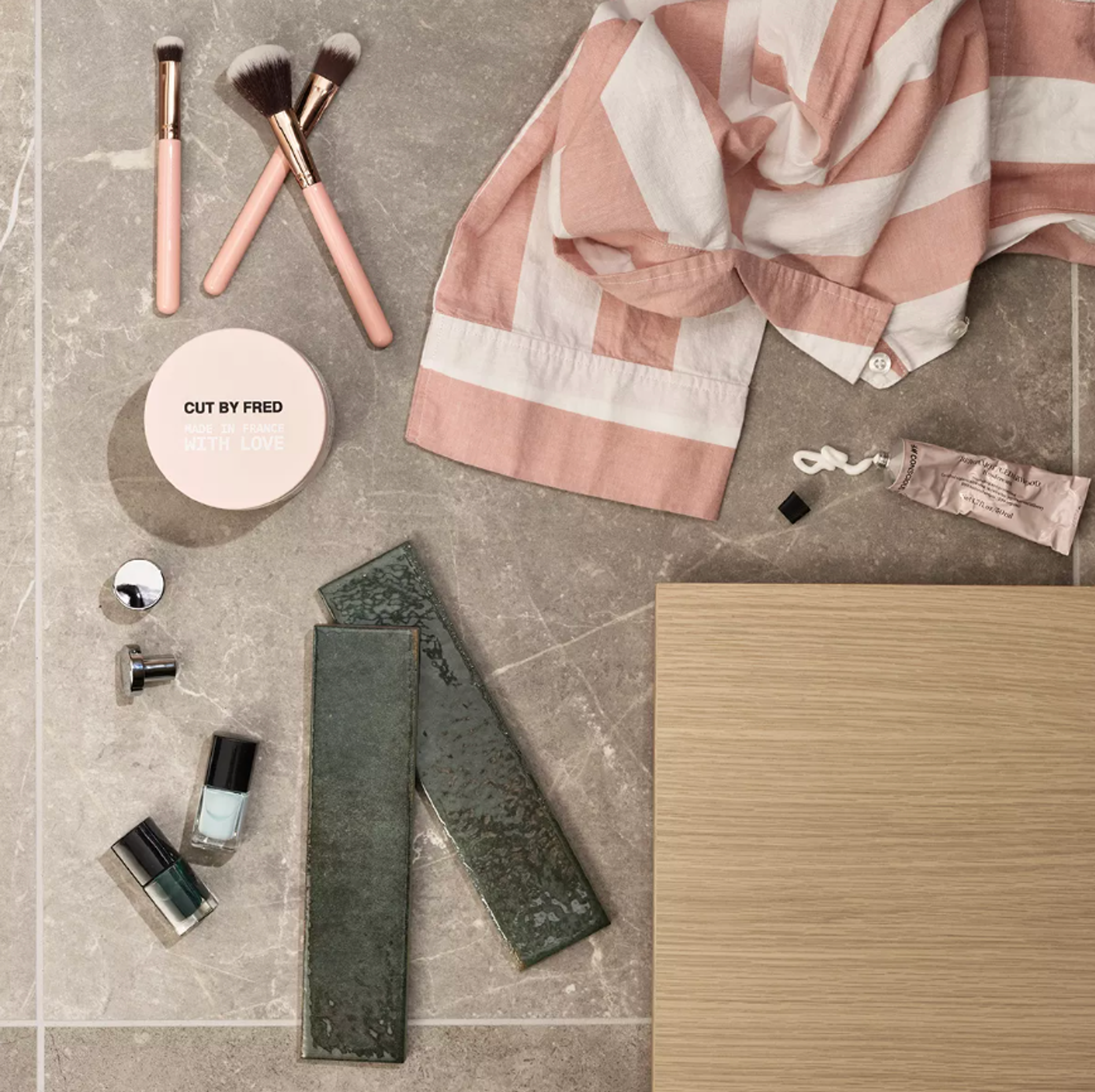 Flat lay of various cosmetics, a pink striped towel, two green tiles, and a wood sample on a grey stone surface.