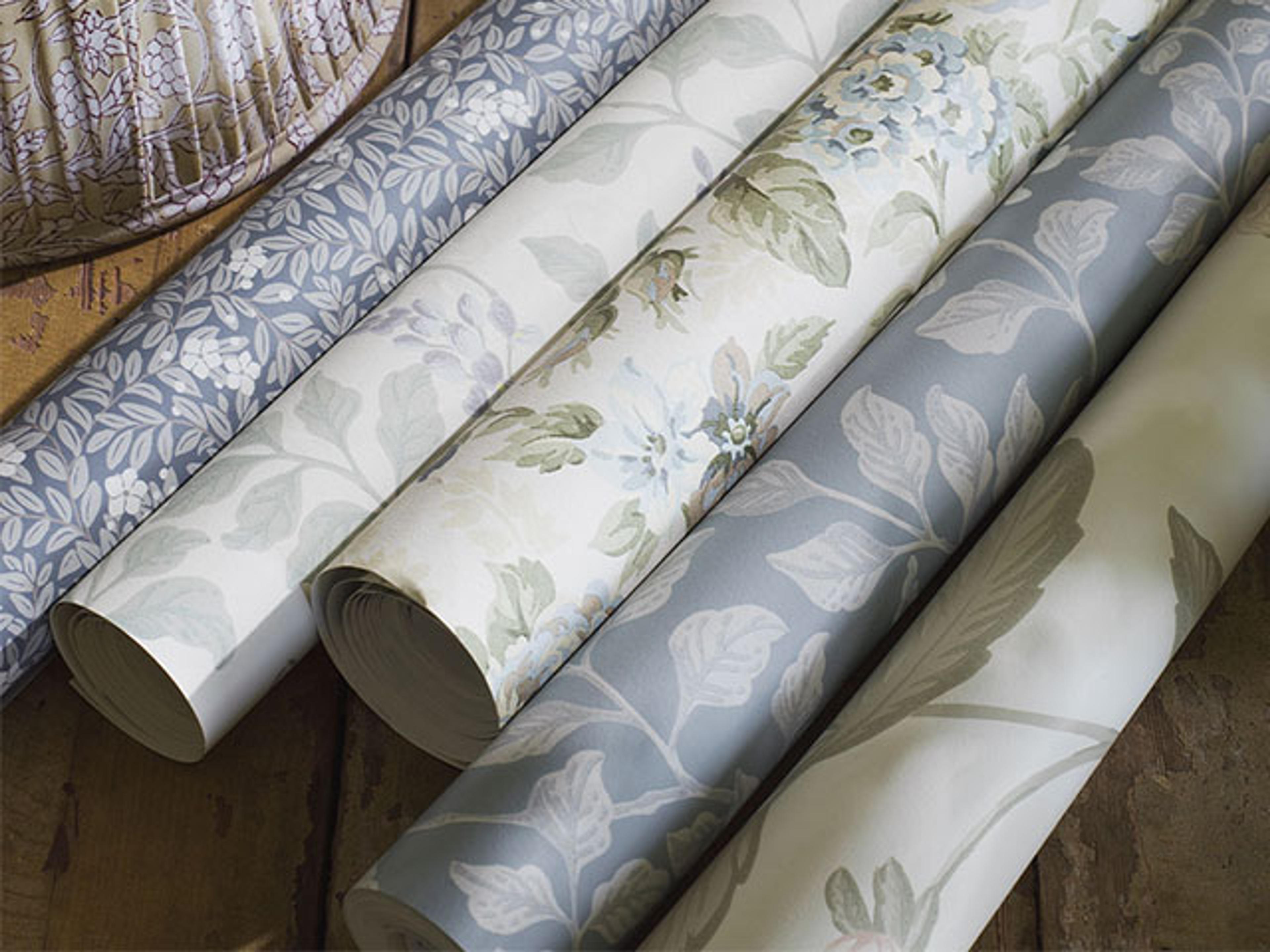 Rolls of wallpaper with various floral and botanical patterns in muted blue, green, and cream tones.