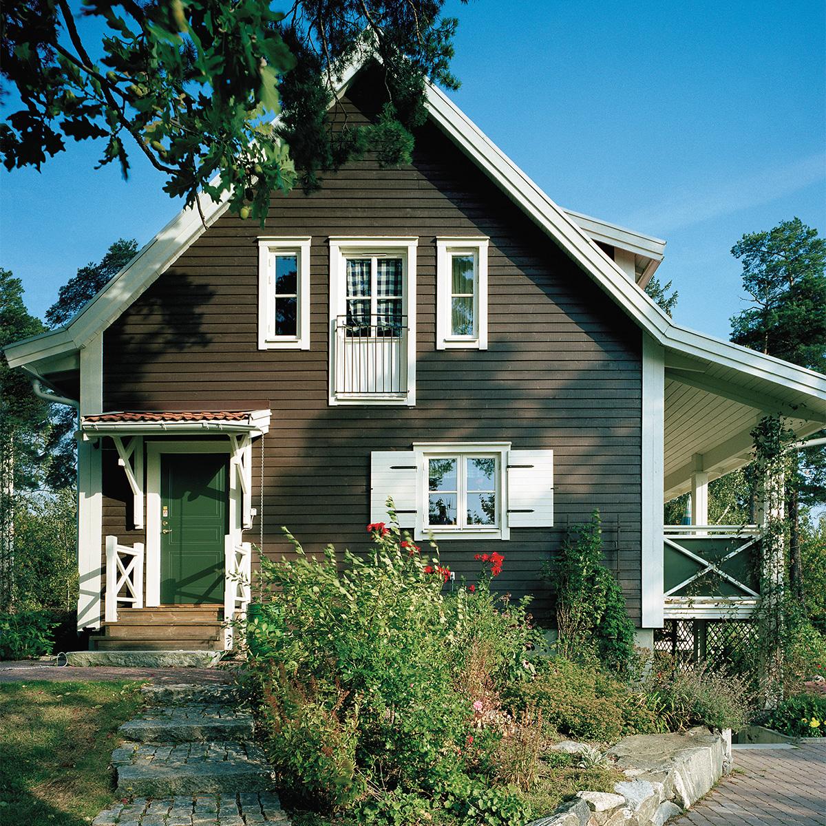 A dark brown two-story house with white trim, green door, and white shutters, set in a lush garden.