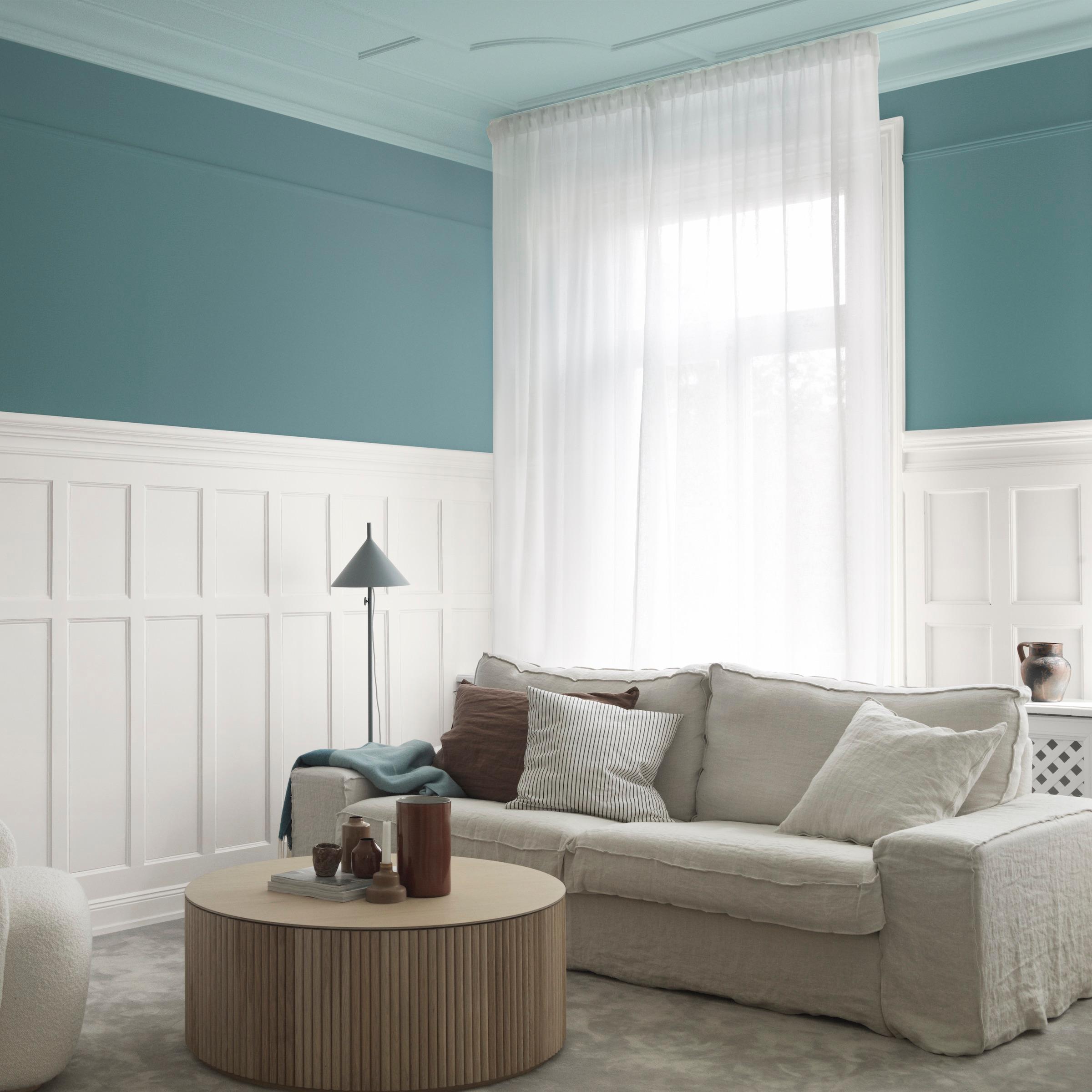 A living room with teal and white paneled walls, a beige sofa, sheer white curtains, and a round slatted wood coffee table.