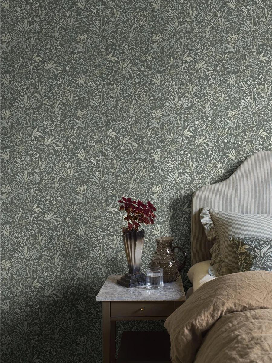 A bedroom with dark green wallpaper featuring a delicate light green foliage pattern, a nightstand with a vase of red leaves, and a partial view of a bed.