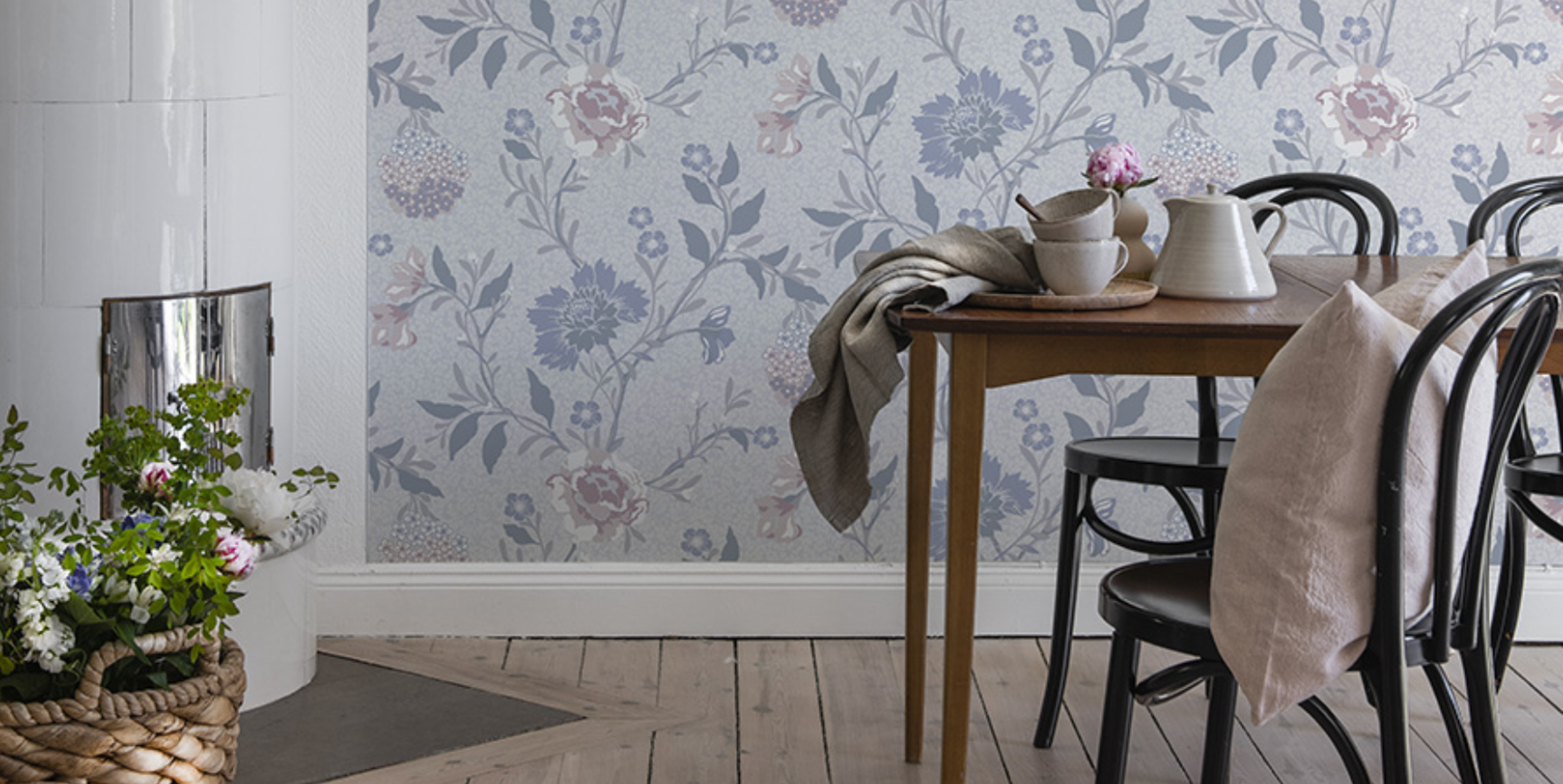 A cozy dining space with light blue floral wallpaper, a wooden table, black chairs, and a white tiled stove.