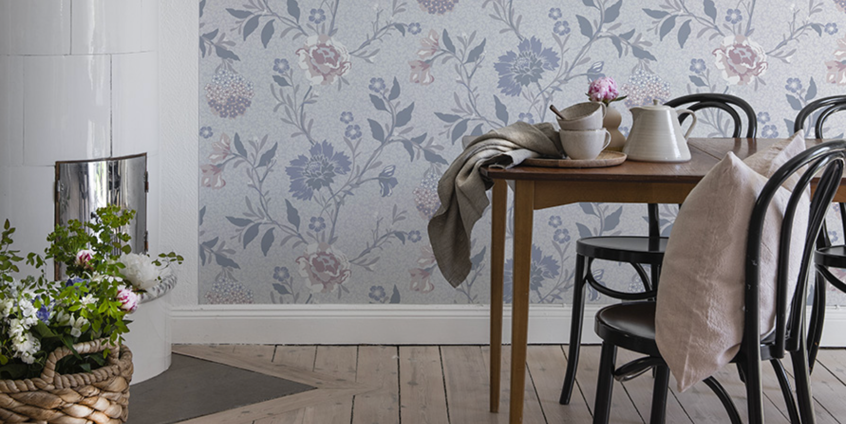 A cozy dining space with light blue floral wallpaper, a wooden table, black chairs, and a white tiled stove.