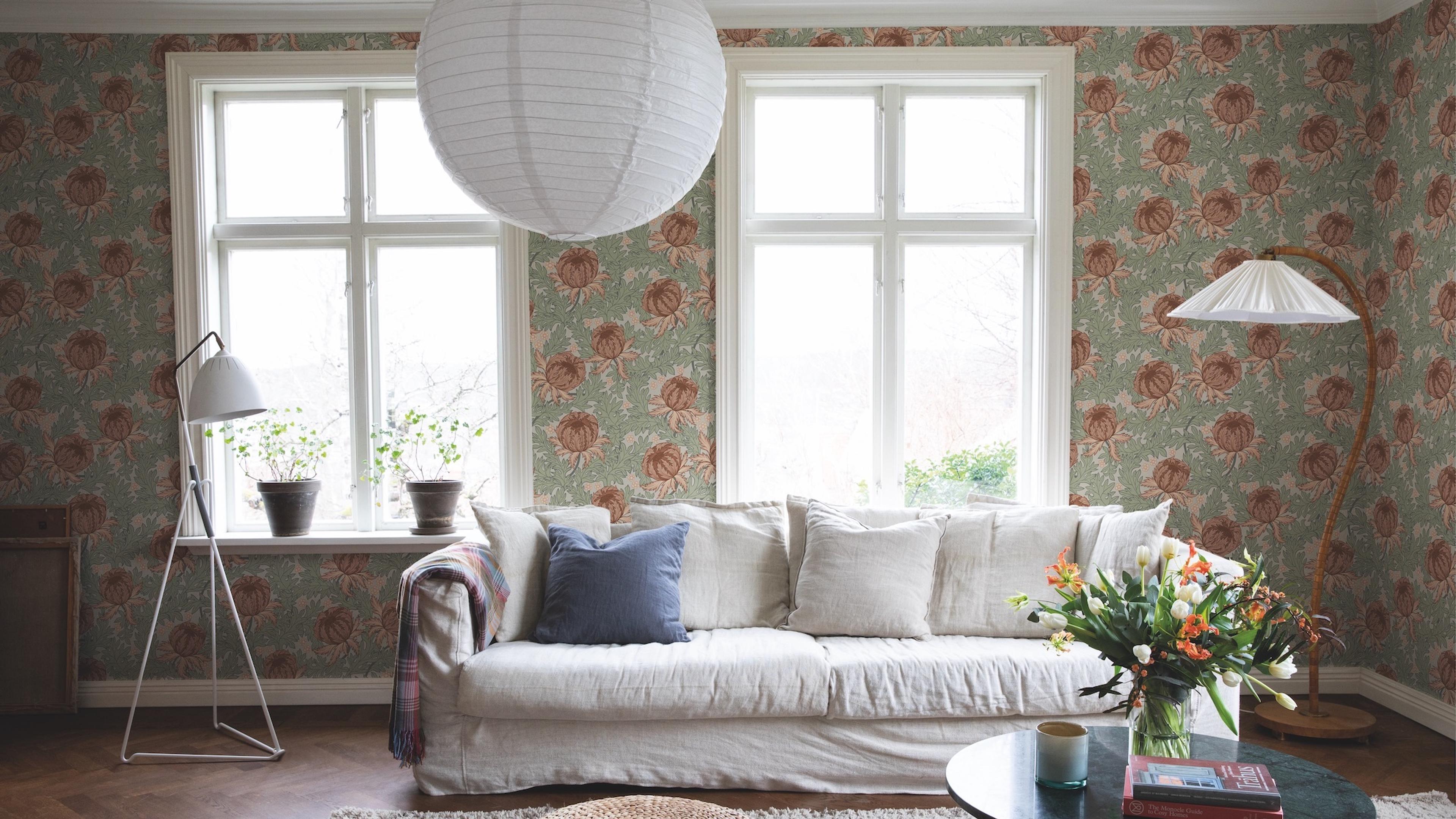 A living room with sage green wallpaper featuring large reddish-brown floral patterns, a white slipcovered sofa, and two windows.