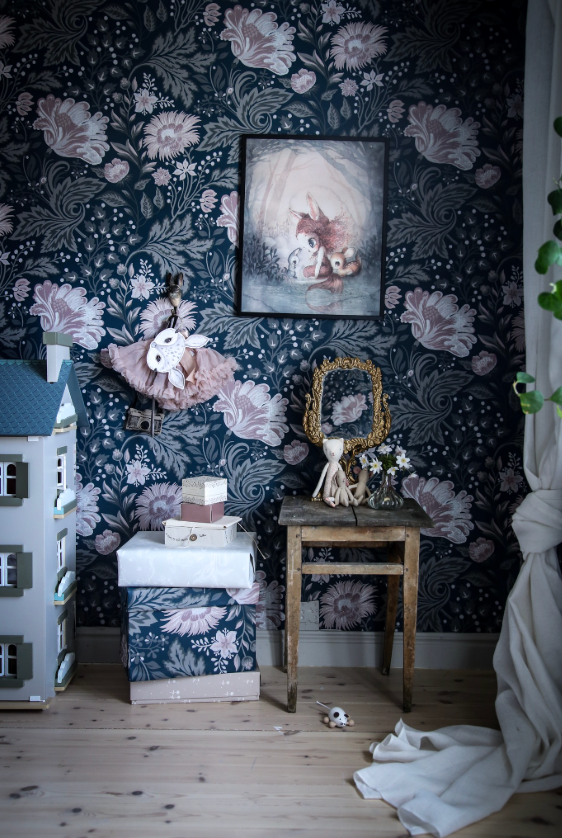 A cozy, whimsical room corner with dark floral wallpaper, a dollhouse, framed art, and decorative items.