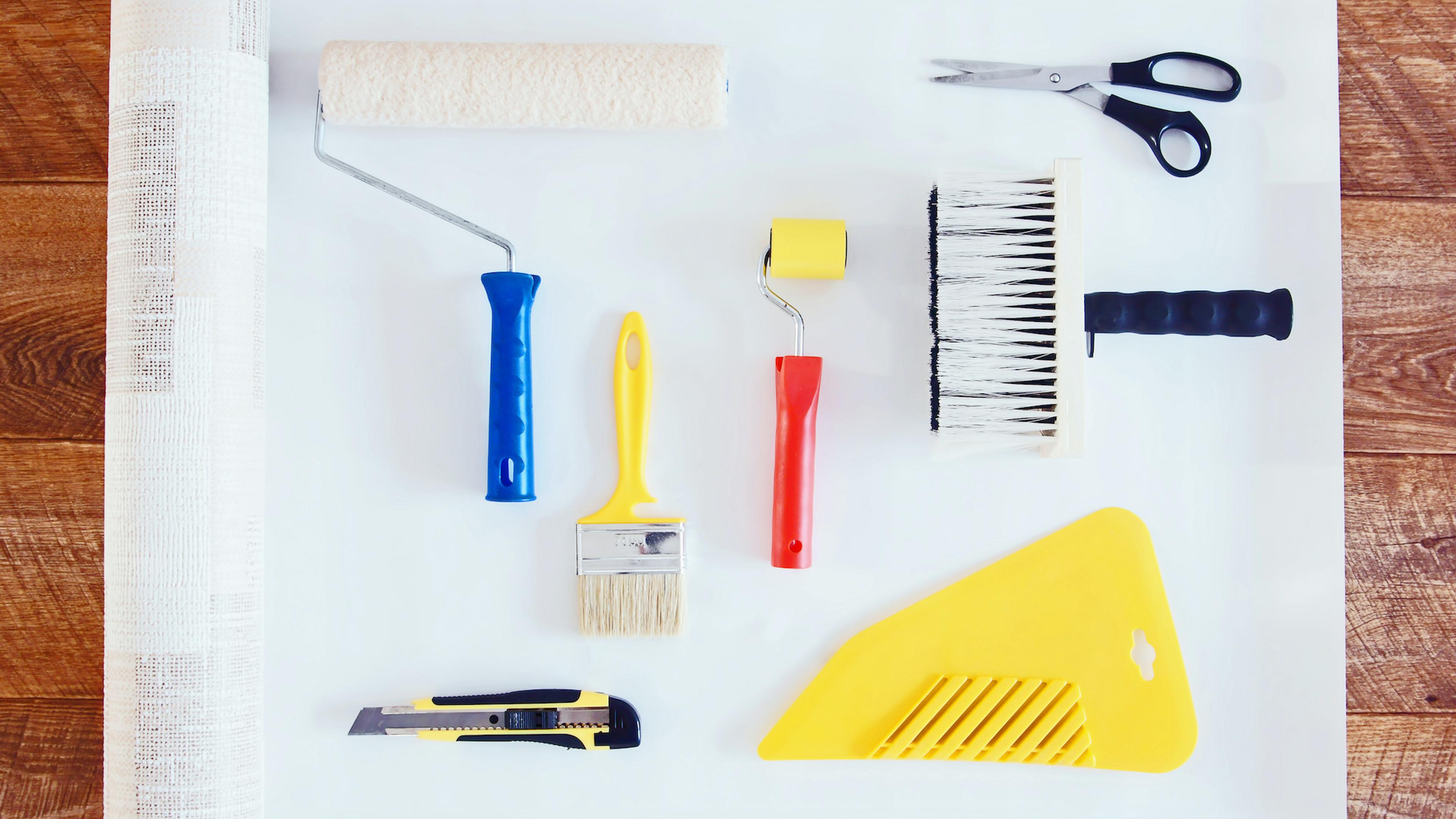 Flat lay of wallpapering tools including a roll of wallpaper, rollers, brushes, scissors, a utility knife, and a smoother.