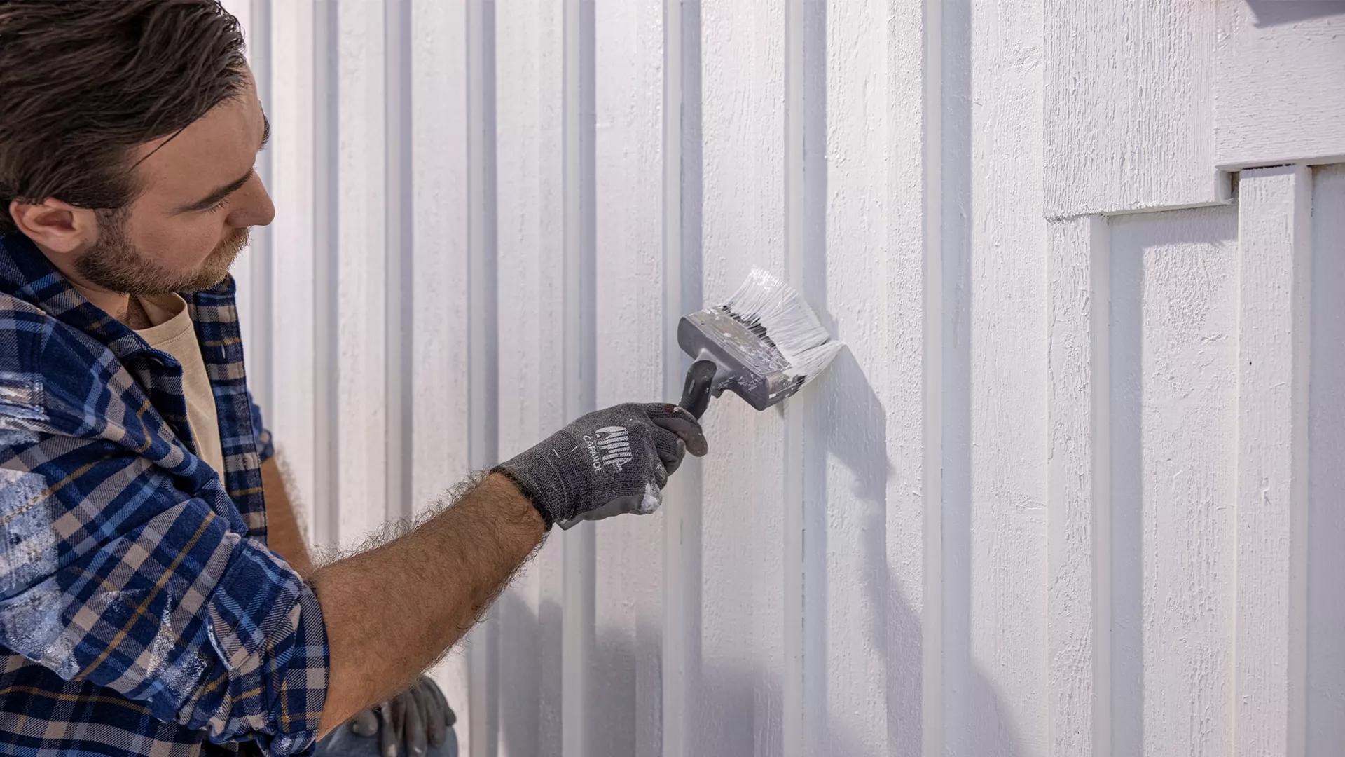 A man in a plaid shirt and gloves paints a white wooden wall with a large brush.