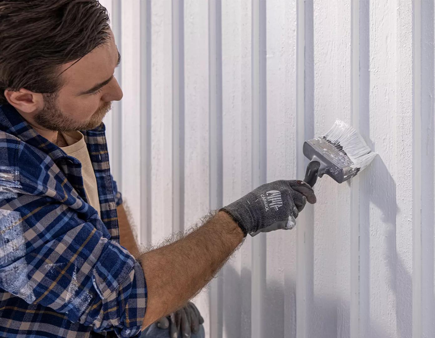 A man in a plaid shirt and gloves paints a white wooden wall with a large brush.