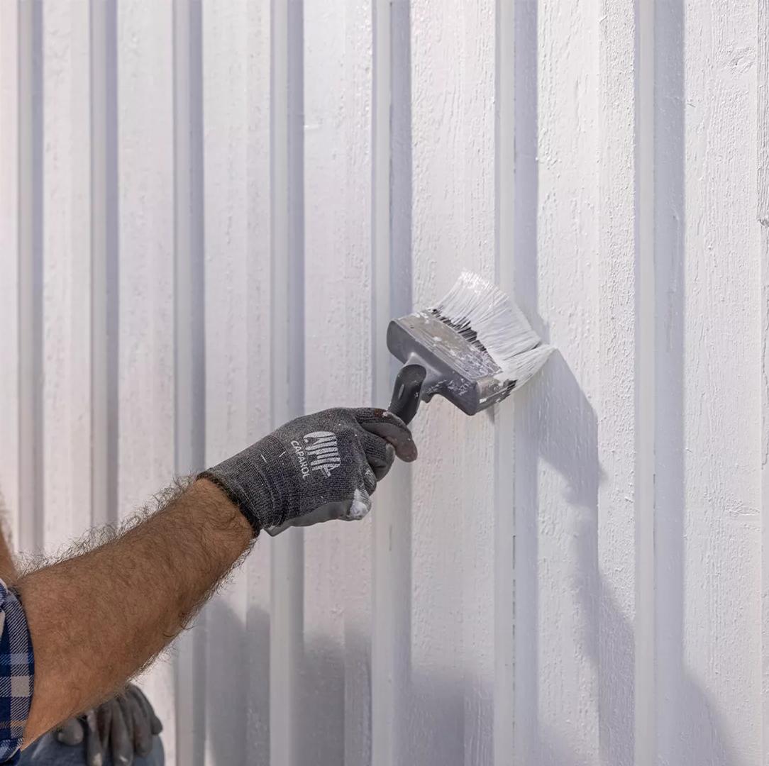 A man in a plaid shirt painting a white wooden wall with a large brush.