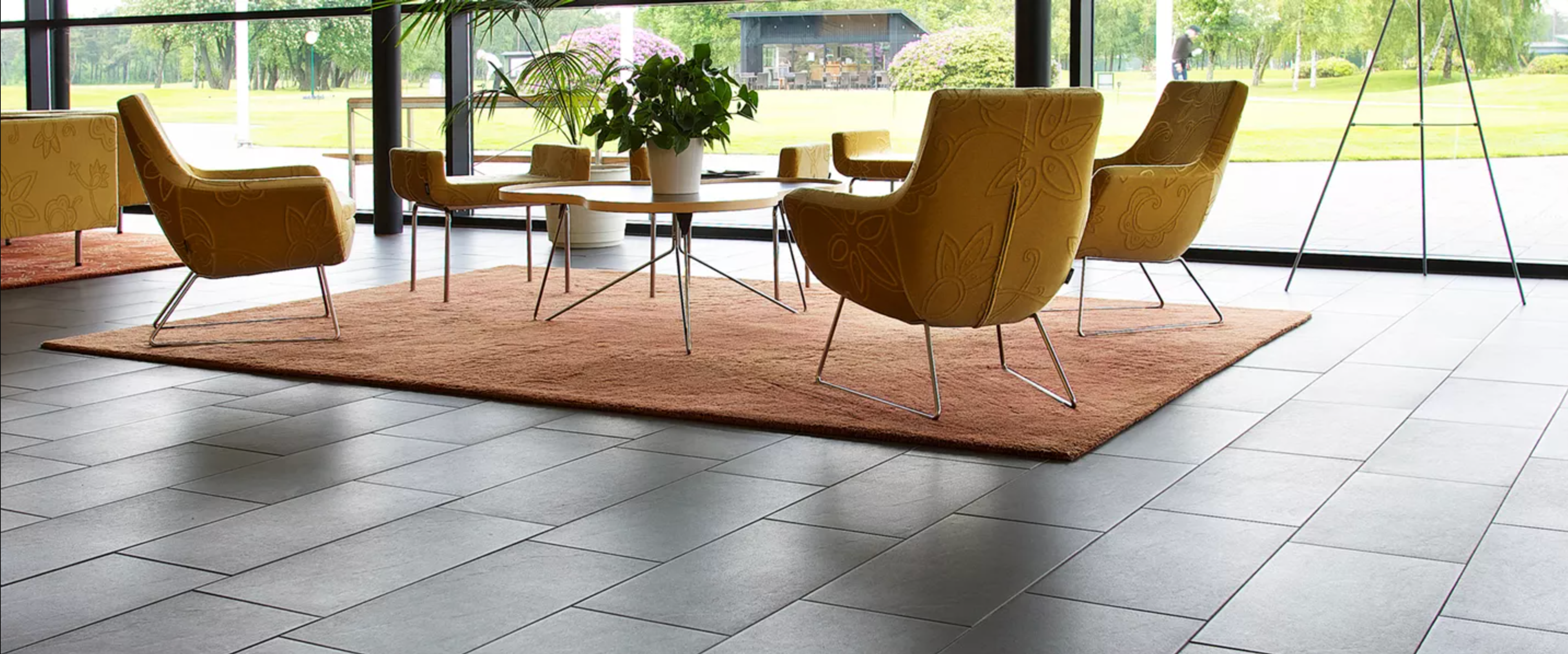 A modern waiting area with dark grey tiled flooring, a brown rug, and mustard-yellow armchairs by large windows.