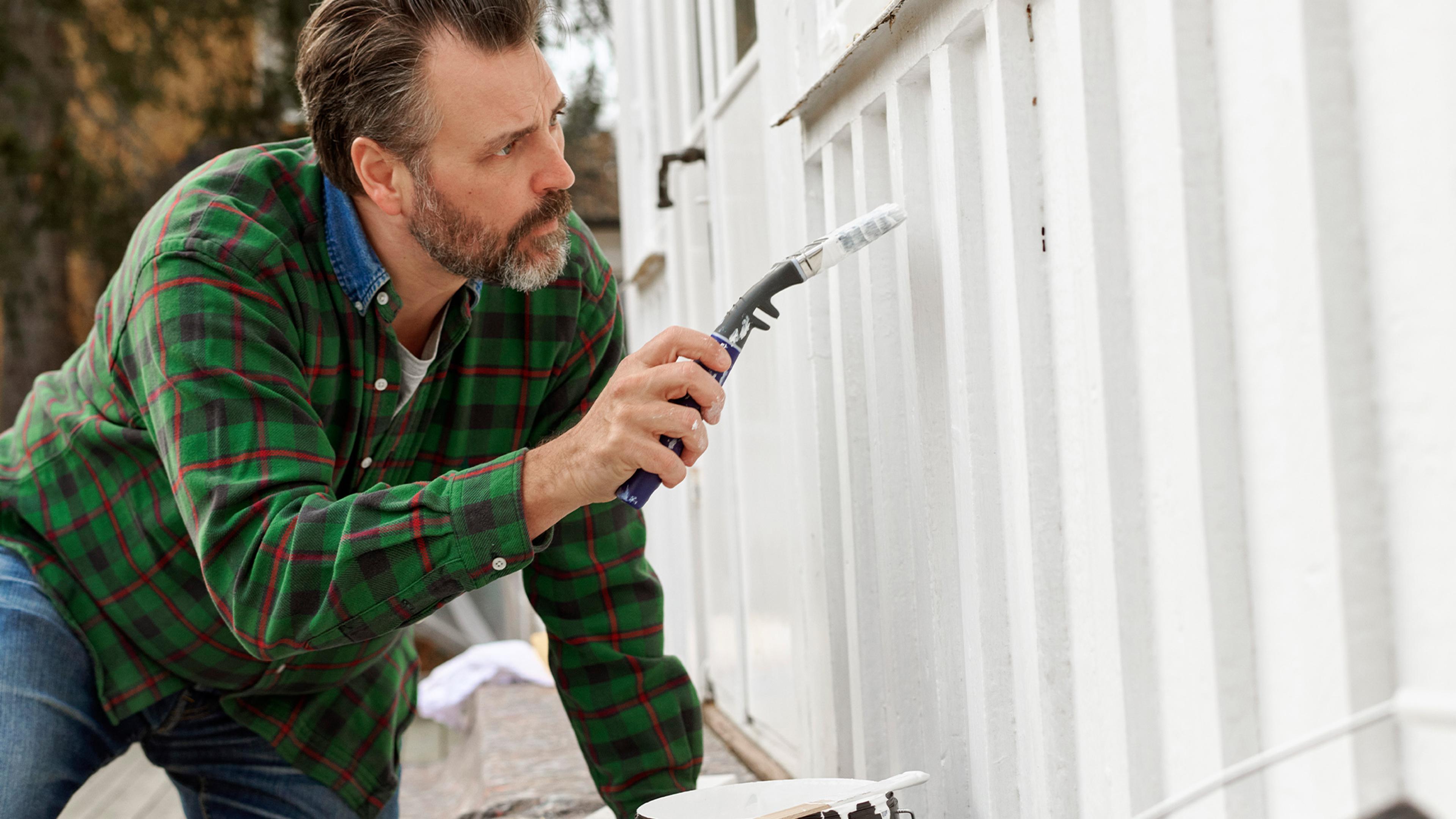 A man in a green plaid shirt paints a white wall.