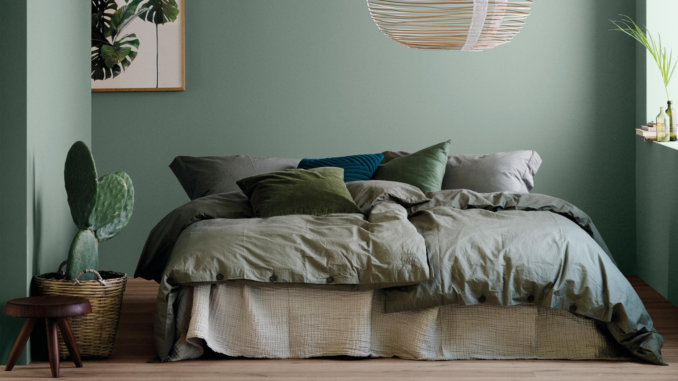 A bedroom with sage green walls, olive green linen bedding, a potted cactus, and natural wood accents.