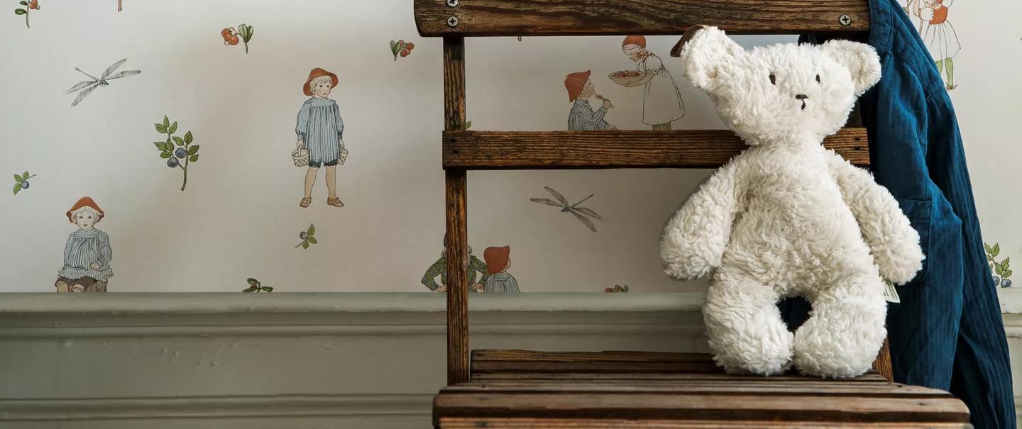 A white teddy bear sits on a wooden stool in front of whimsical wallpaper with children and nature illustrations.