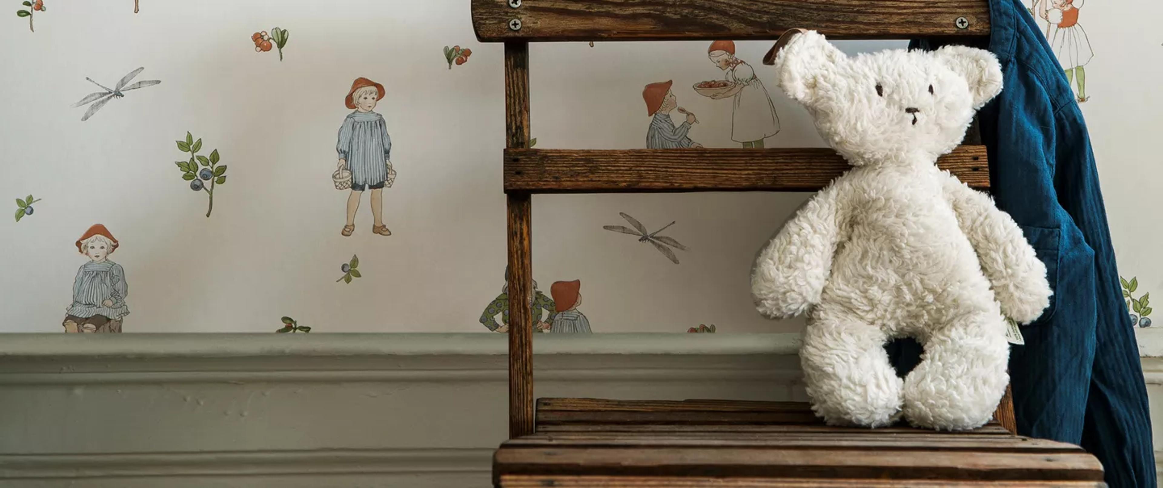 A white teddy bear sits on a wooden stool in front of whimsical wallpaper with children and nature illustrations.