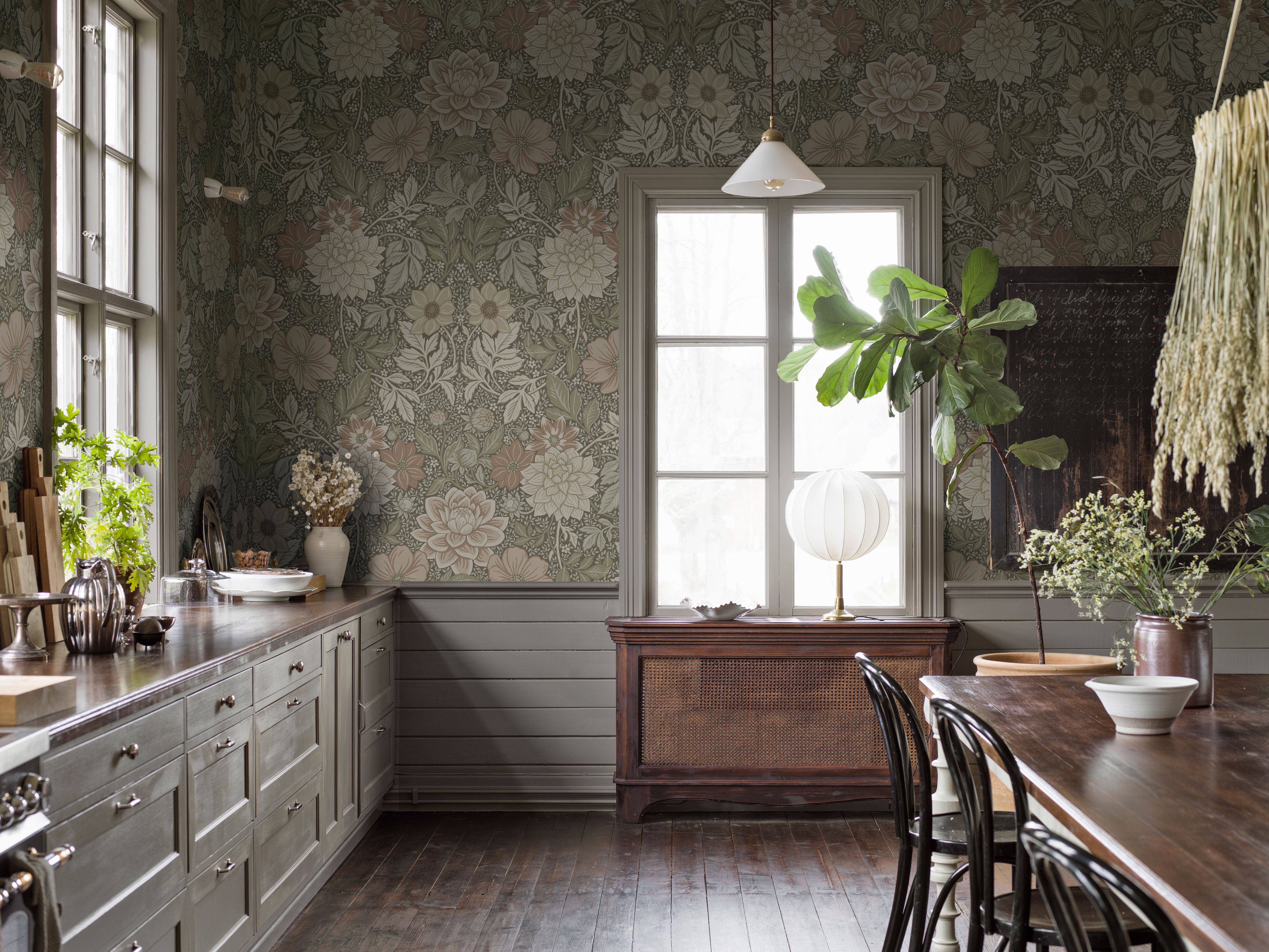 A rustic kitchen and dining room with sage green floral wallpaper, gray cabinets, wooden floors, and natural decor.