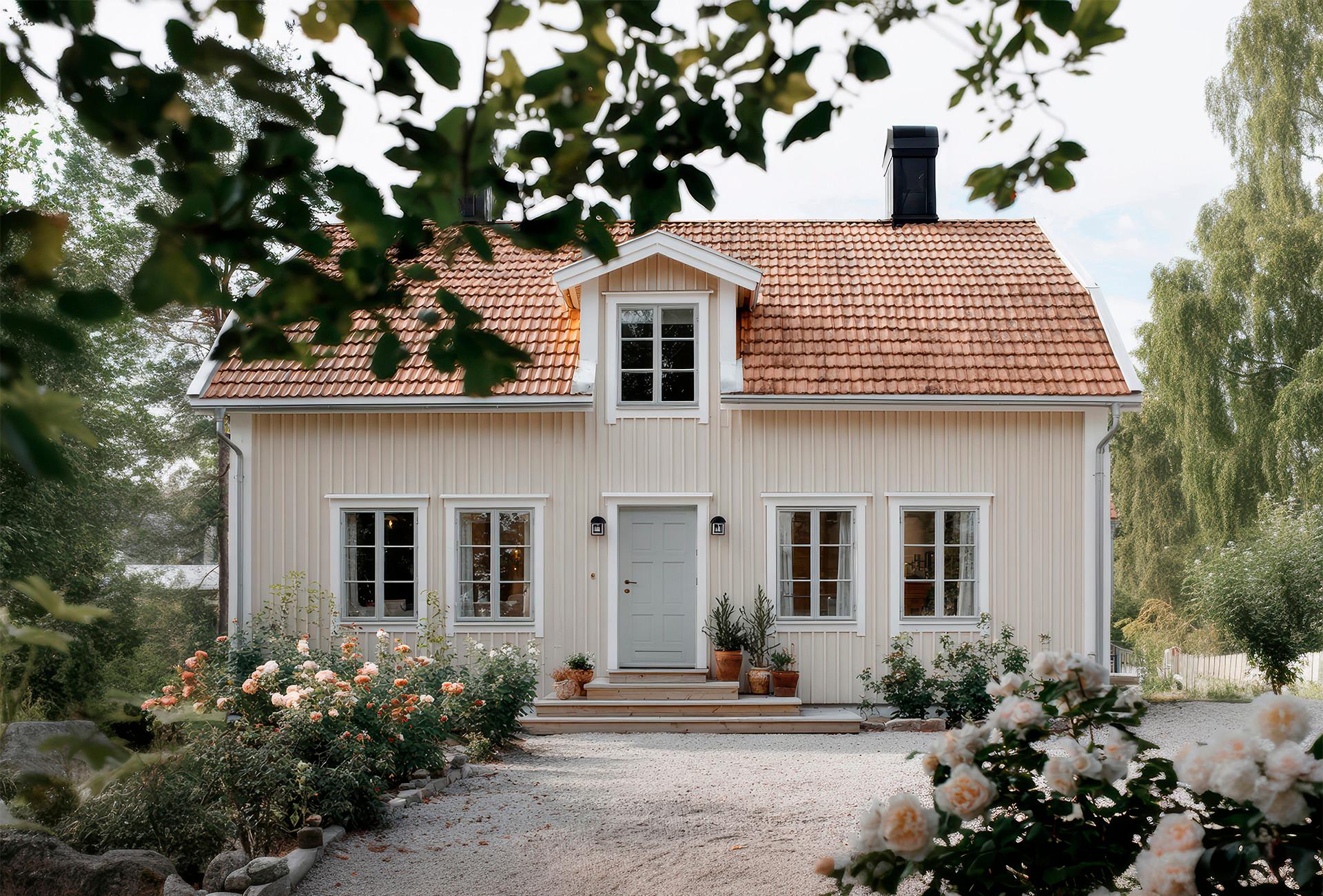 A light yellow house with a red tiled roof, gray front door, and a garden of blooming rose bushes.