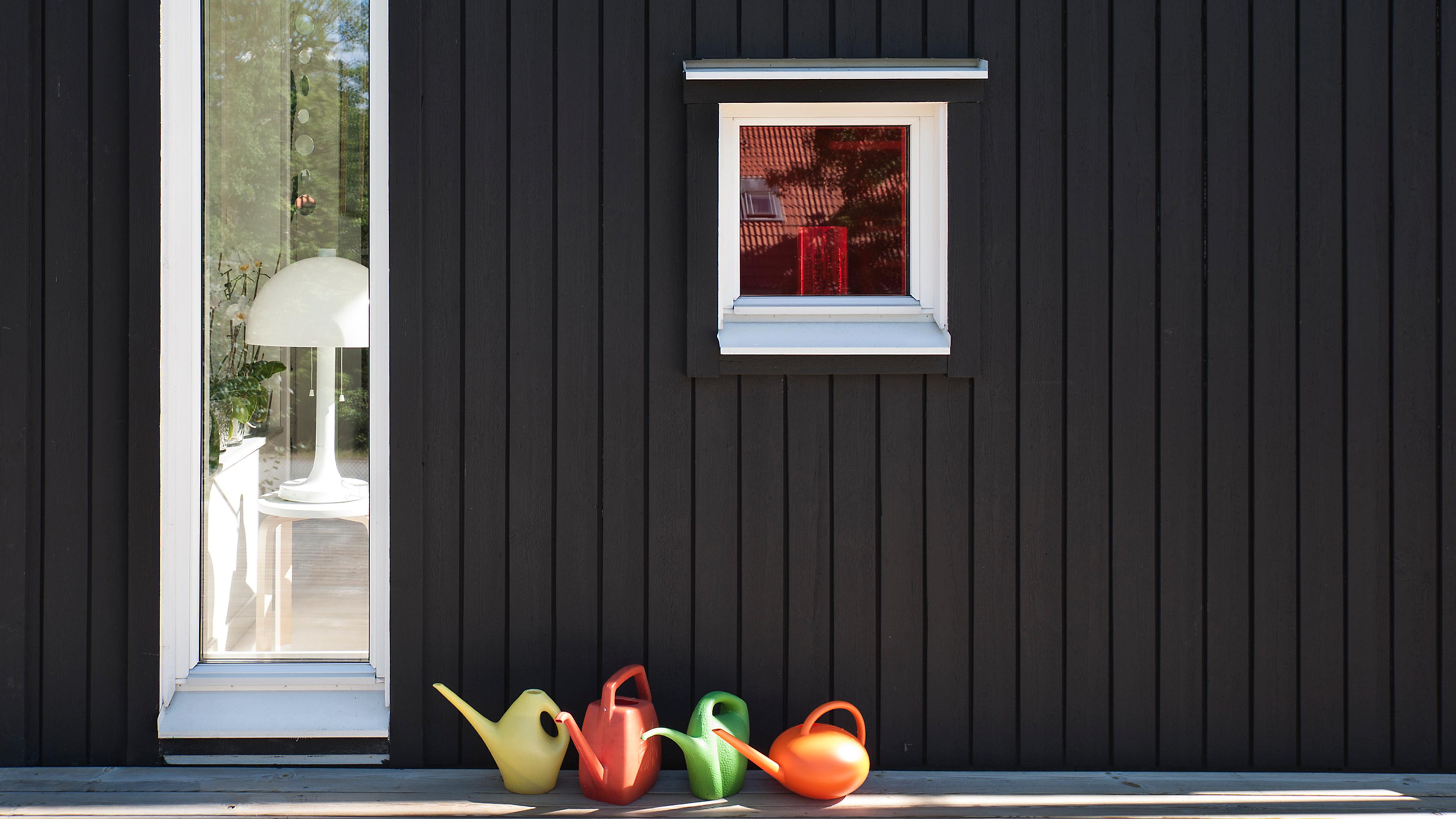 A dark paneled wall with a tall window showing a white lamp, a smaller red-tinted window, and four colorful watering cans below.
