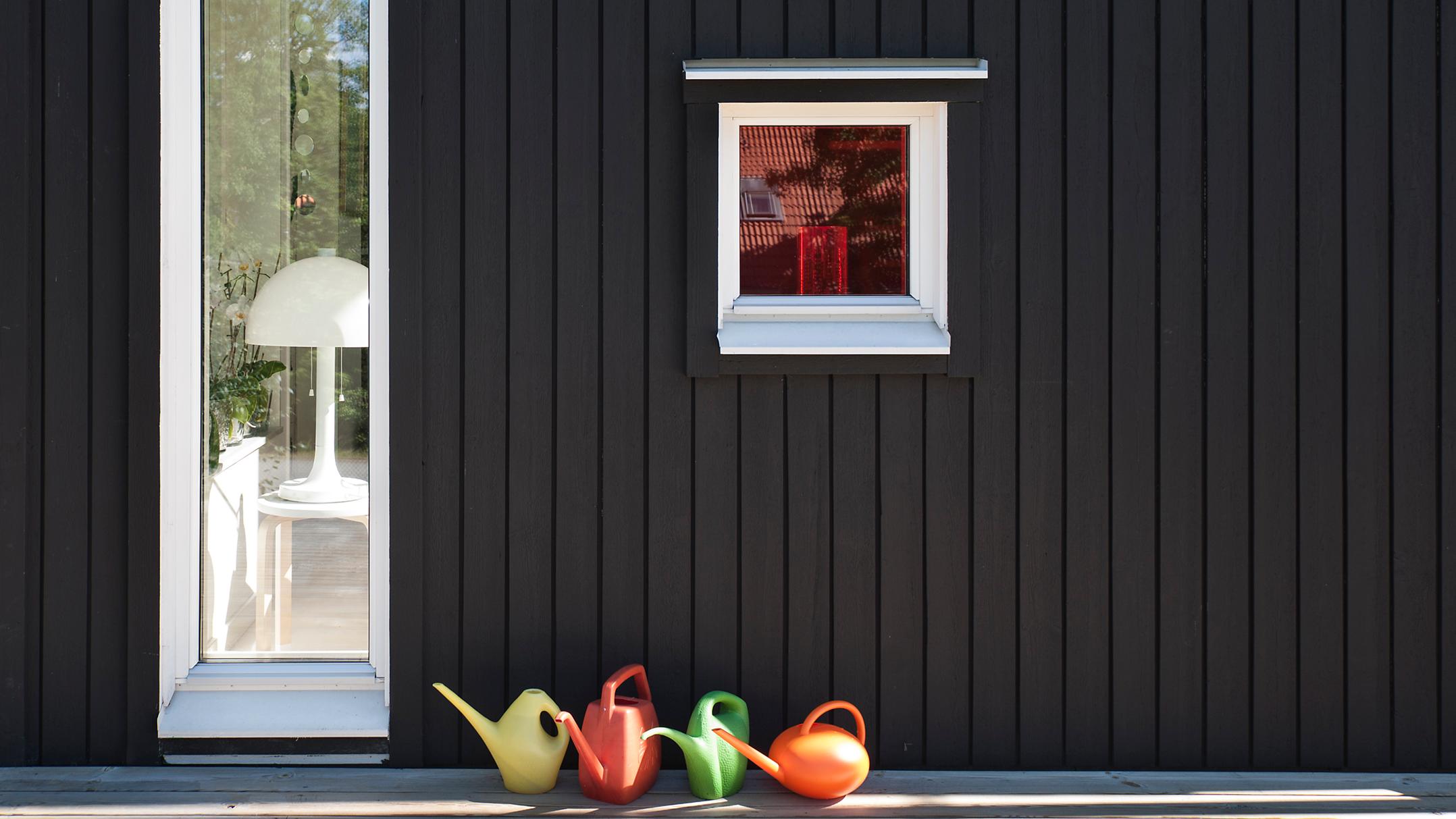 A dark paneled wall with a tall window showing a white lamp, a smaller red-tinted window, and four colorful watering cans below.