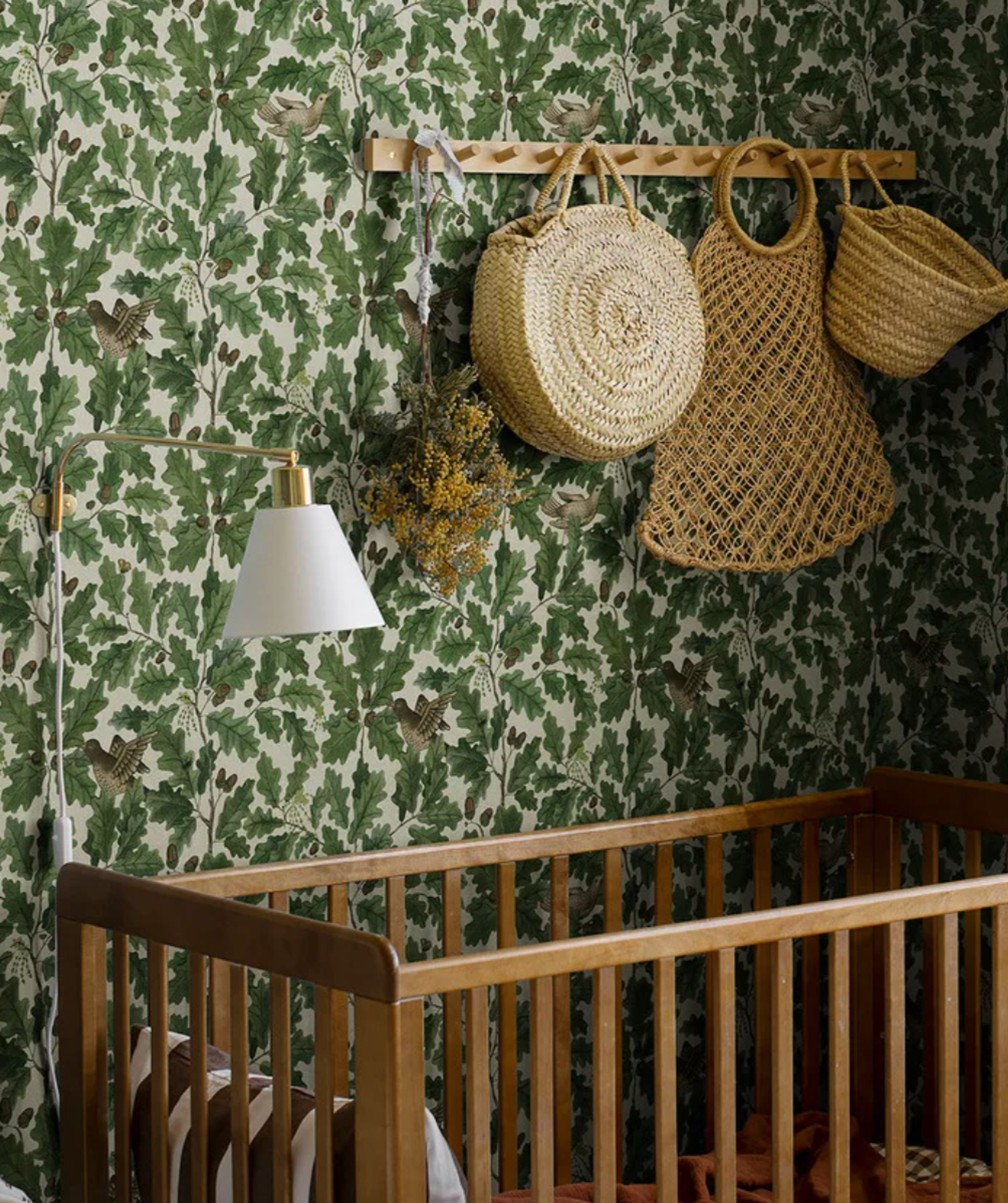 A wooden baby crib sits against a wall covered in green oak leaf and bird pattern wallpaper, with a lamp and woven bags hanging above.