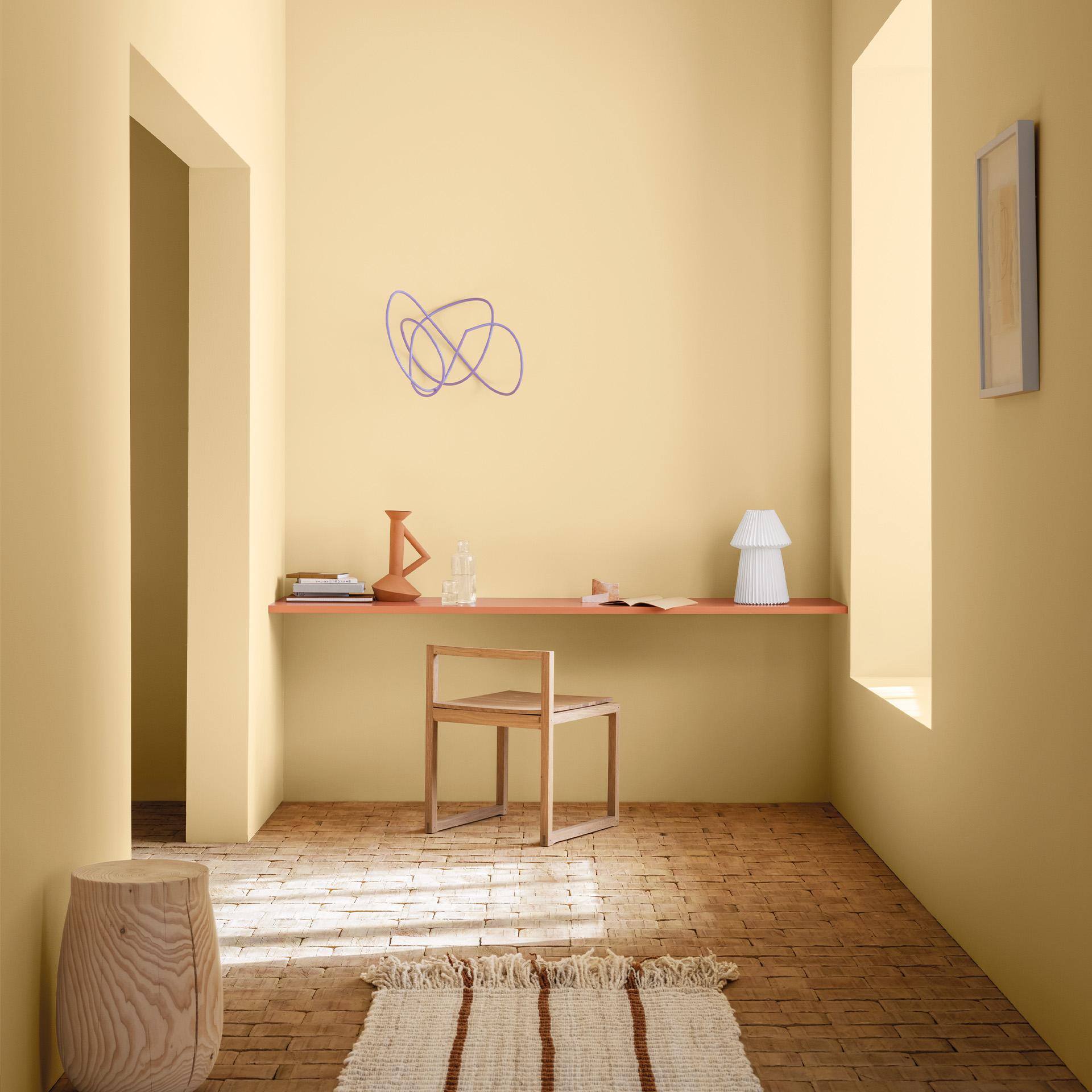 Minimalist home office with pale yellow walls, a floating wooden desk, a simple chair, a wooden stool, and a striped rug on a brick floor.