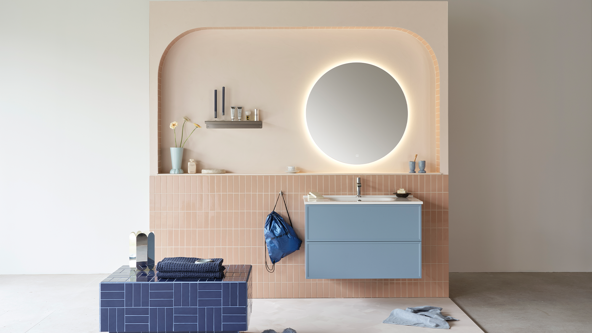 A modern bathroom with a light pink arched wall feature, pink vertical tiles, a backlit round mirror, a blue floating vanity, and a blue tiled bench.