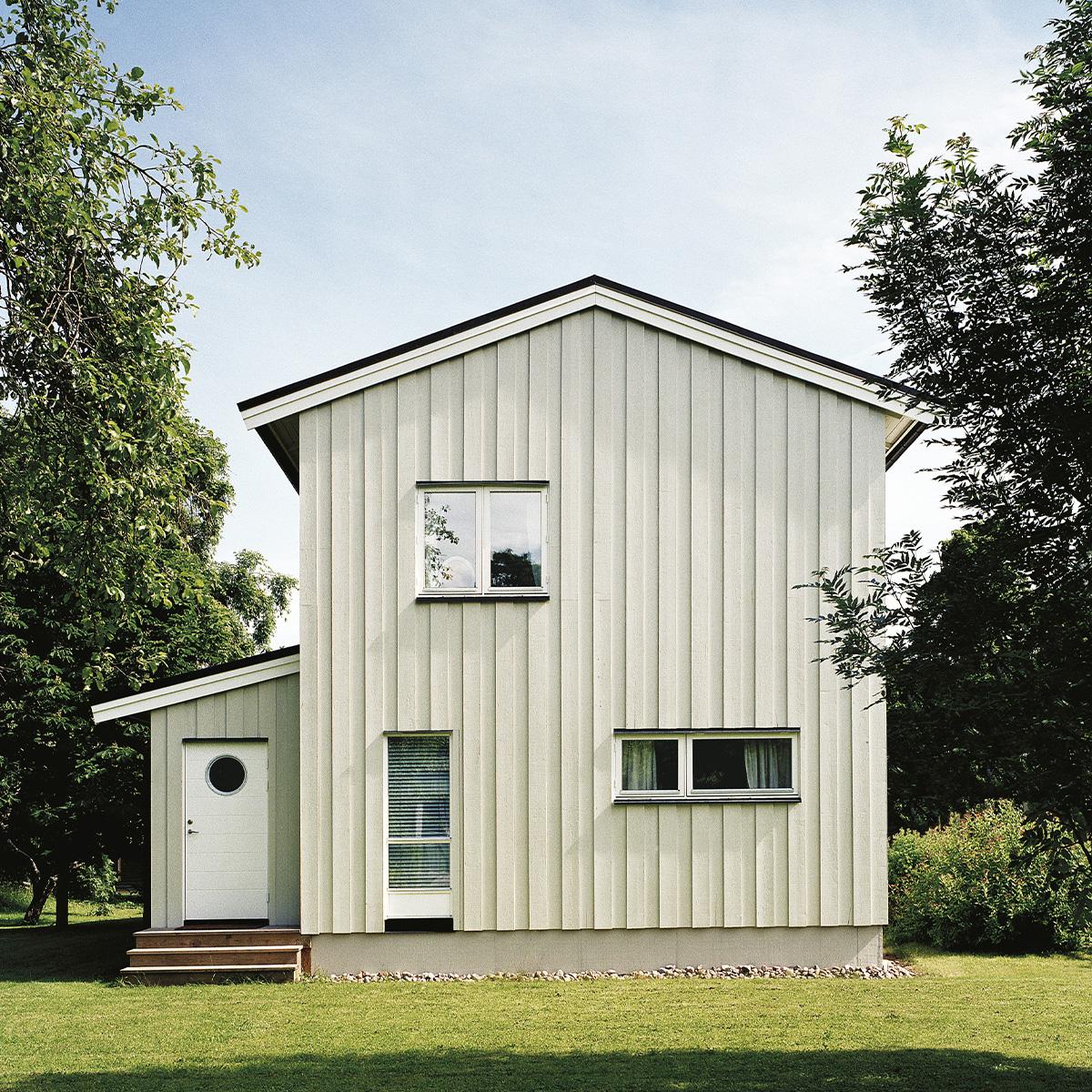 A light green vertical-sided house with a gabled roof and a white door featuring a round window, on a green lawn surrounded by trees.
