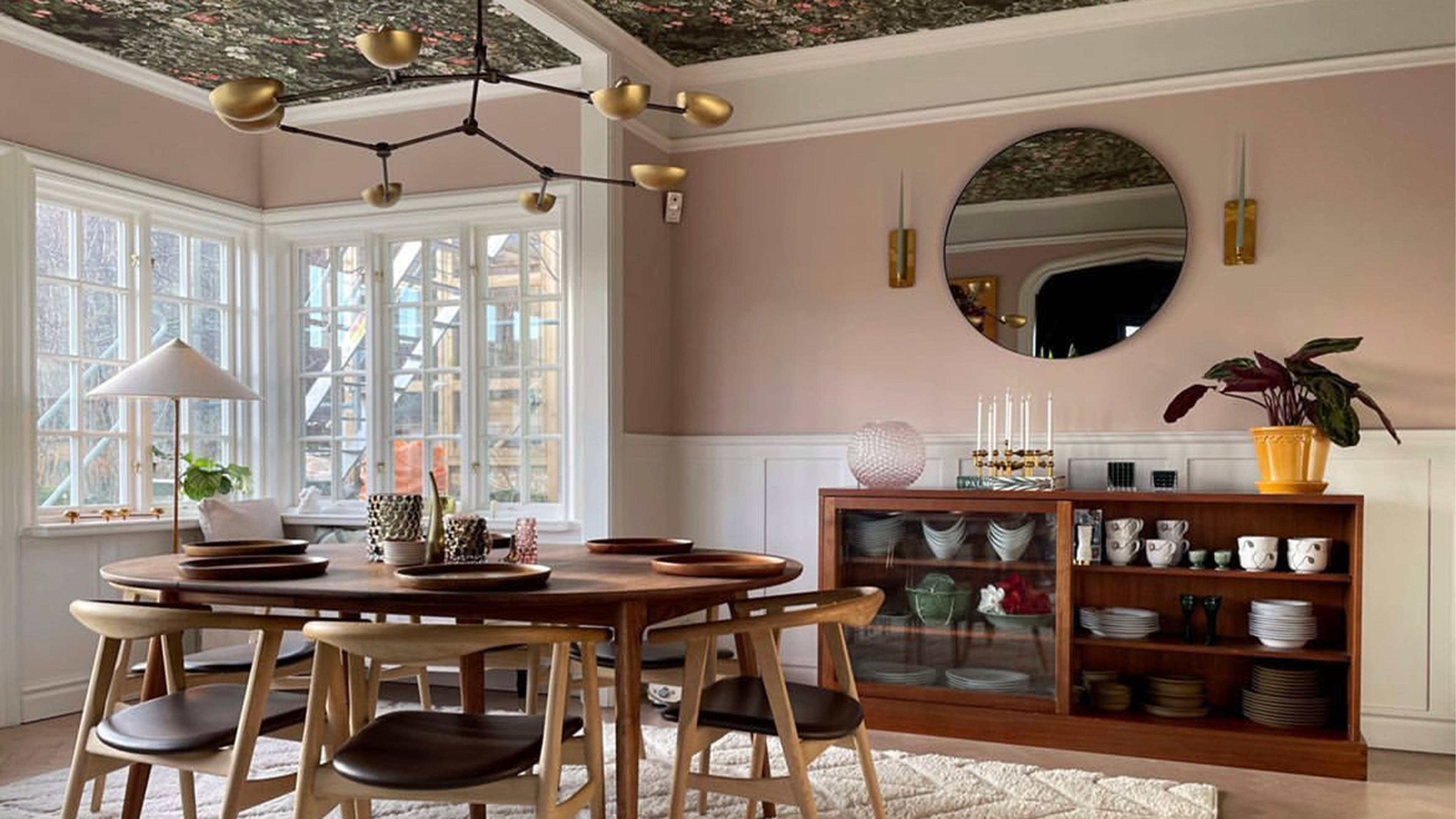 Eclectic dining room with pink walls, a floral ceiling, oval wood table, modern chandelier, wood sideboard, and round mirror.