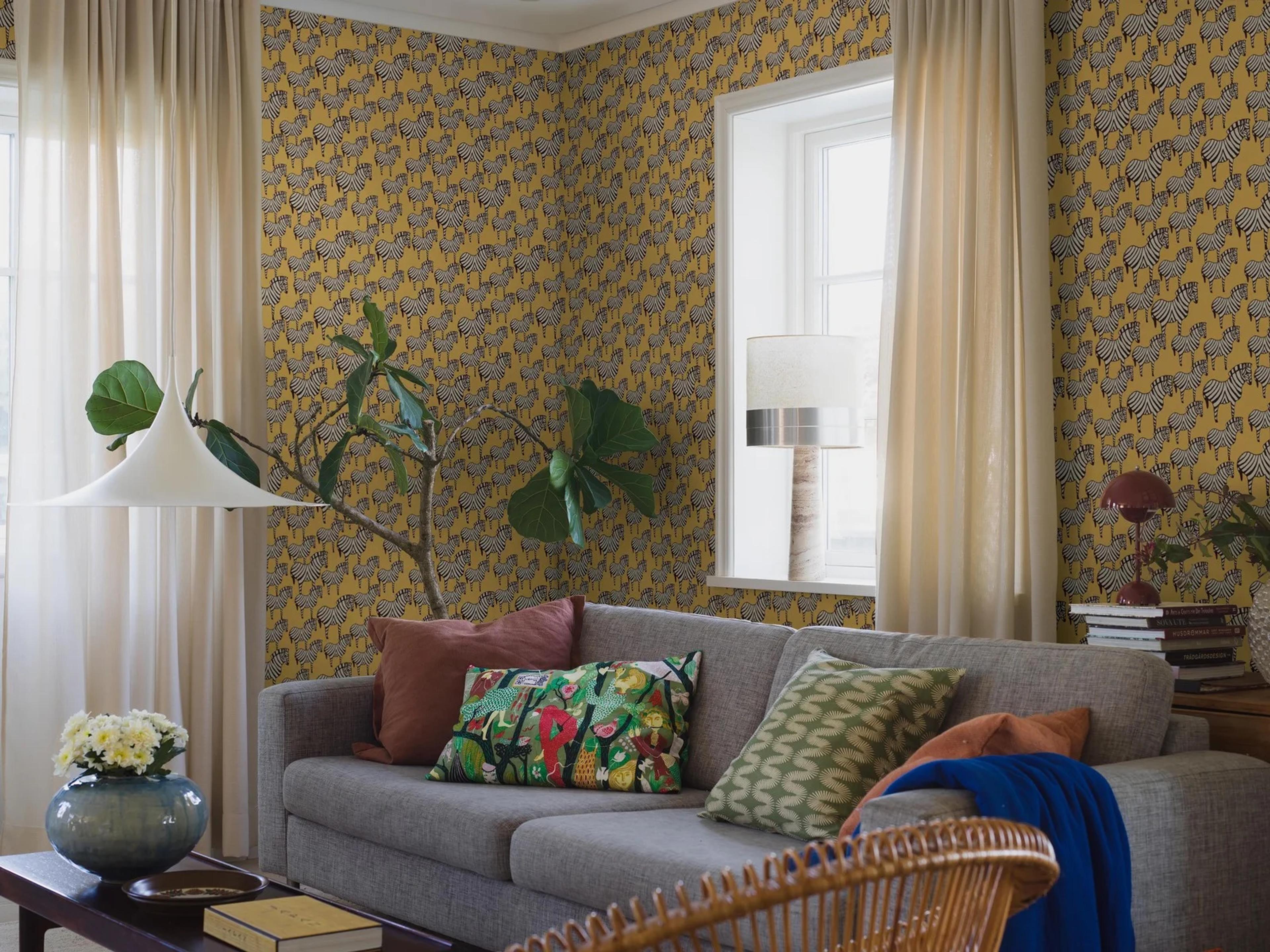 A living room with vibrant yellow zebra-patterned wallpaper, a grey sofa, and light curtains.