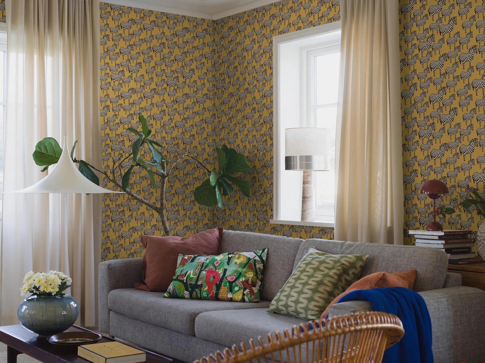 A living room with vibrant yellow zebra-patterned wallpaper, a grey sofa, and light curtains.
