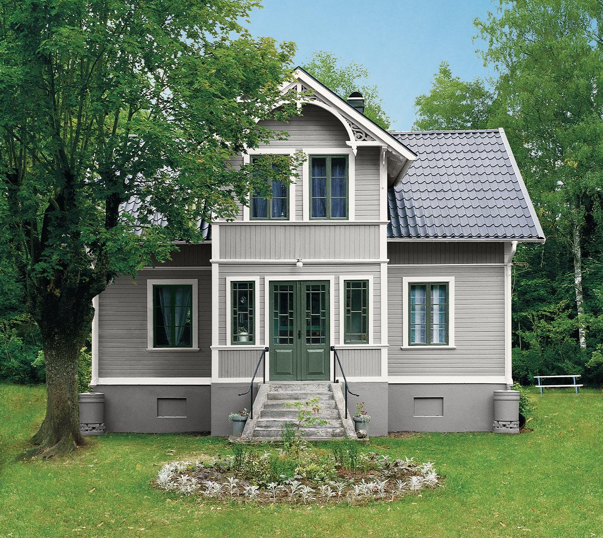 A light gray house with dark green doors and window frames, a dark tiled roof, set in a green lawn with trees.