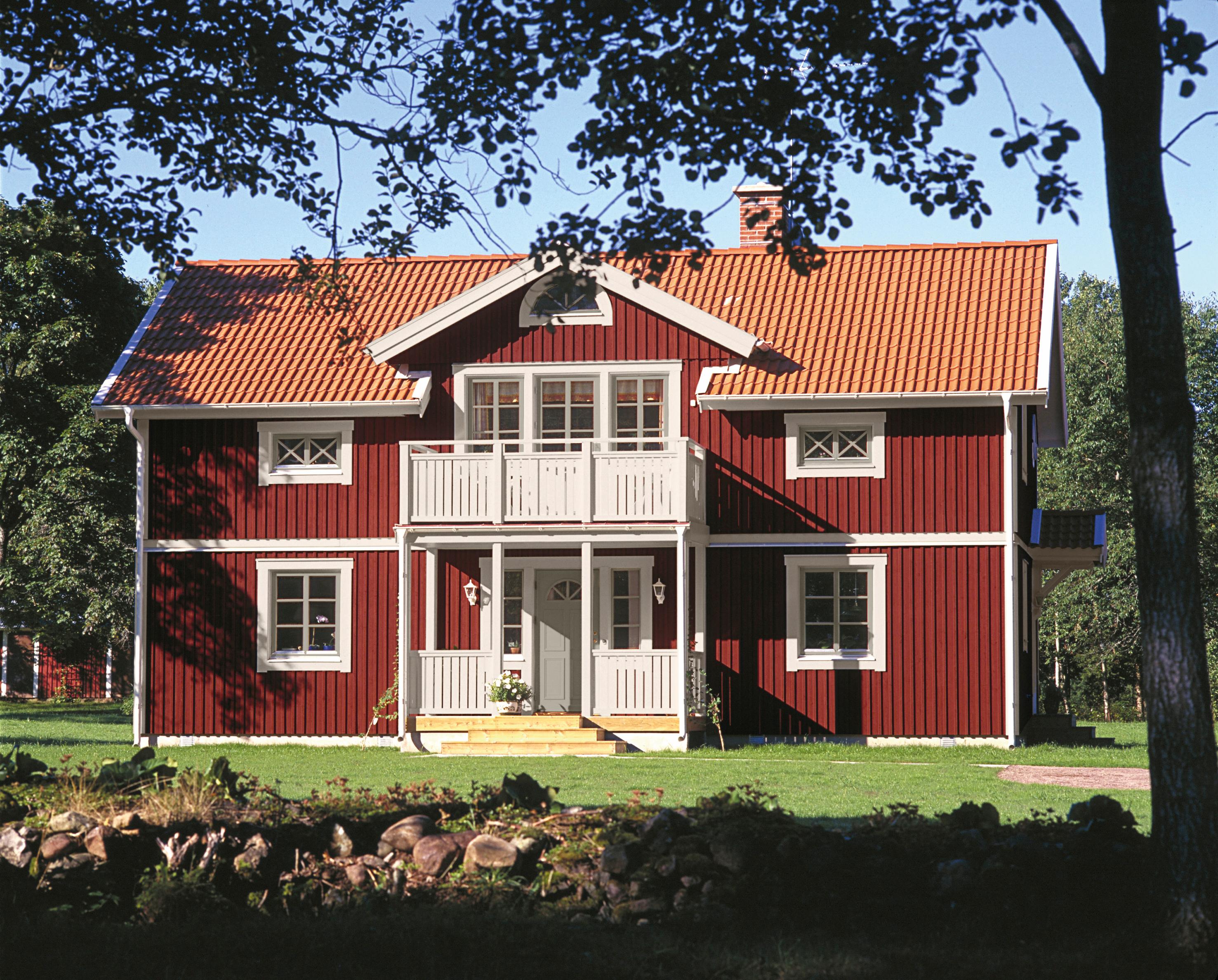 A traditional red house with a terracotta roof, white trim, and a balcony, surrounded by a green lawn and trees.