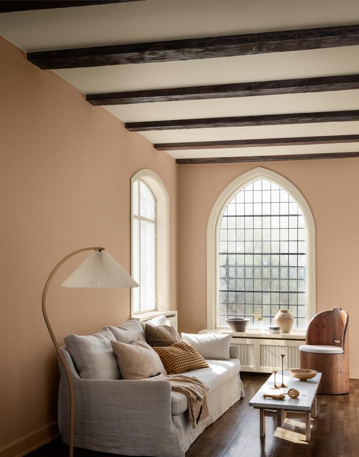 A living room with tan walls, dark exposed ceiling beams, arched windows, a light gray sofa, a floor lamp, and a wooden accent chair.