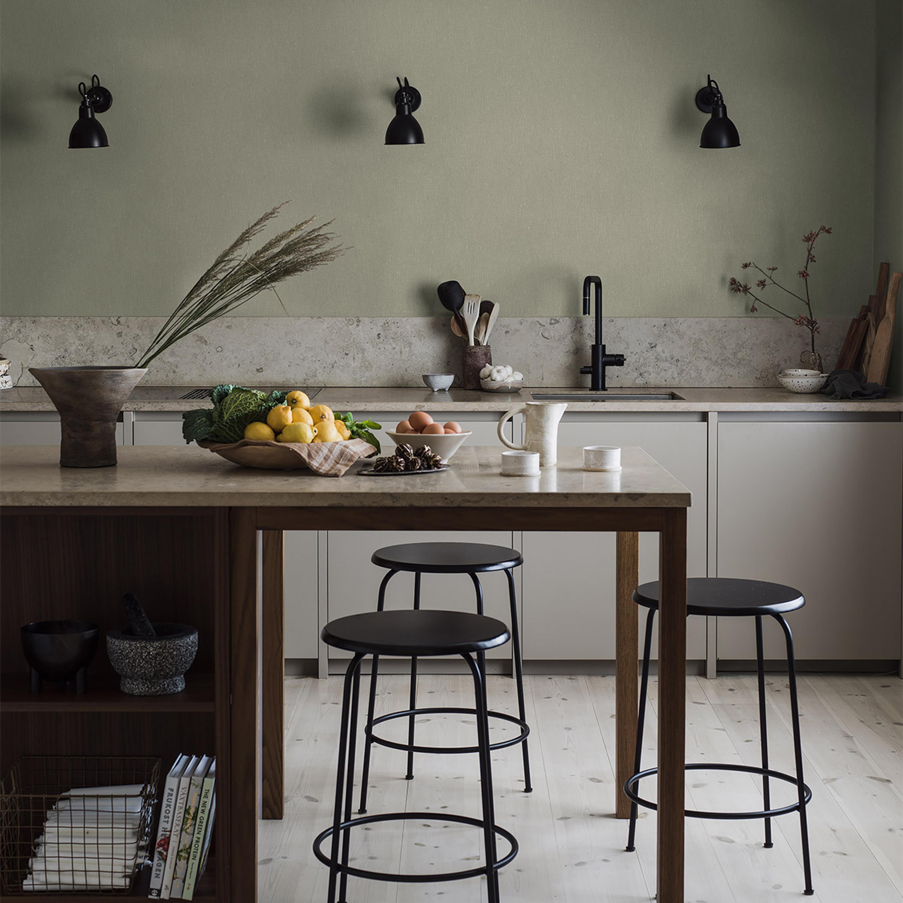 A stylish kitchen with sage green walls, a large island with a wooden bowl of lemons and greens, eggs, and three black bar stools.