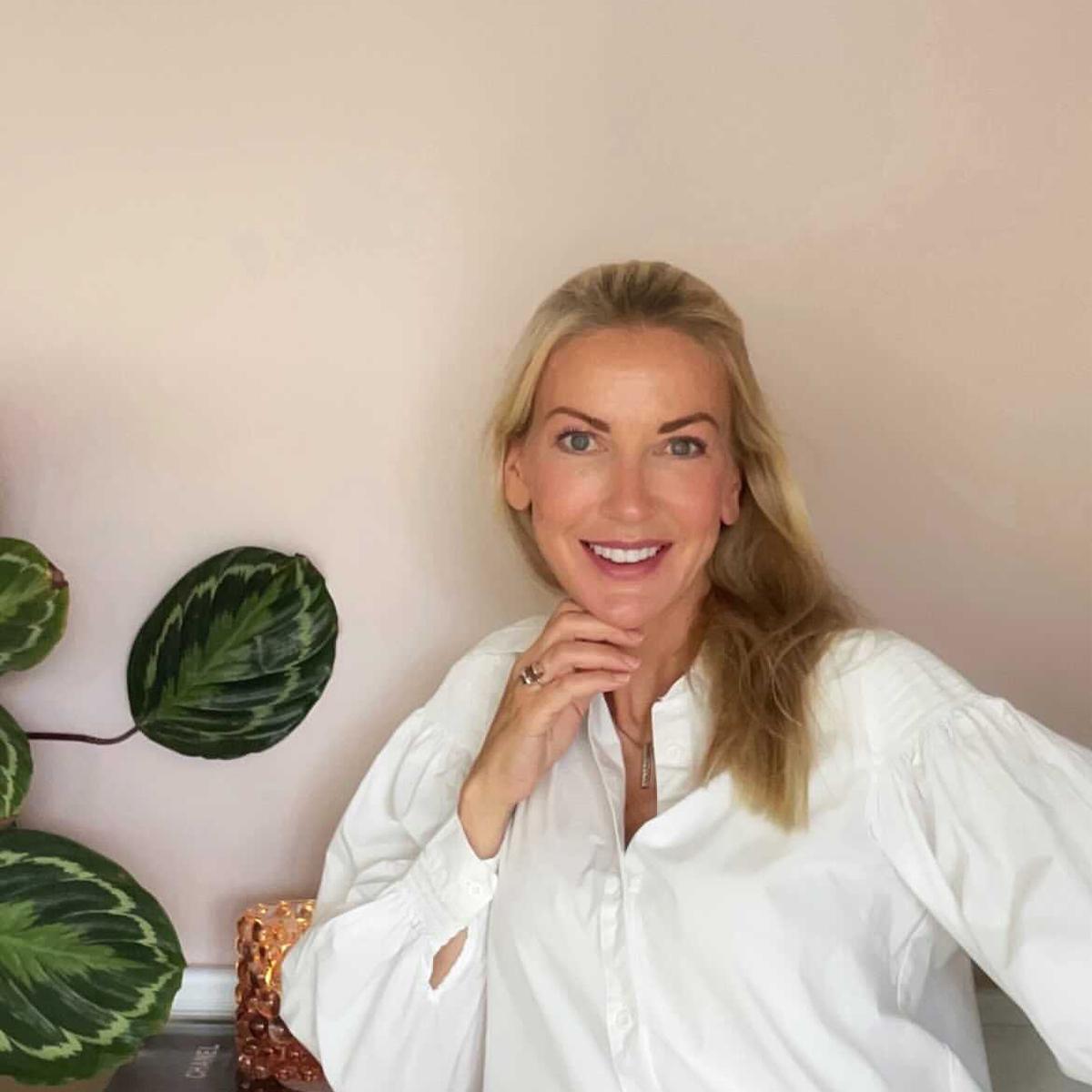 A smiling blonde woman in a white collared shirt rests her chin on her hand, with a leafy green plant on the left.