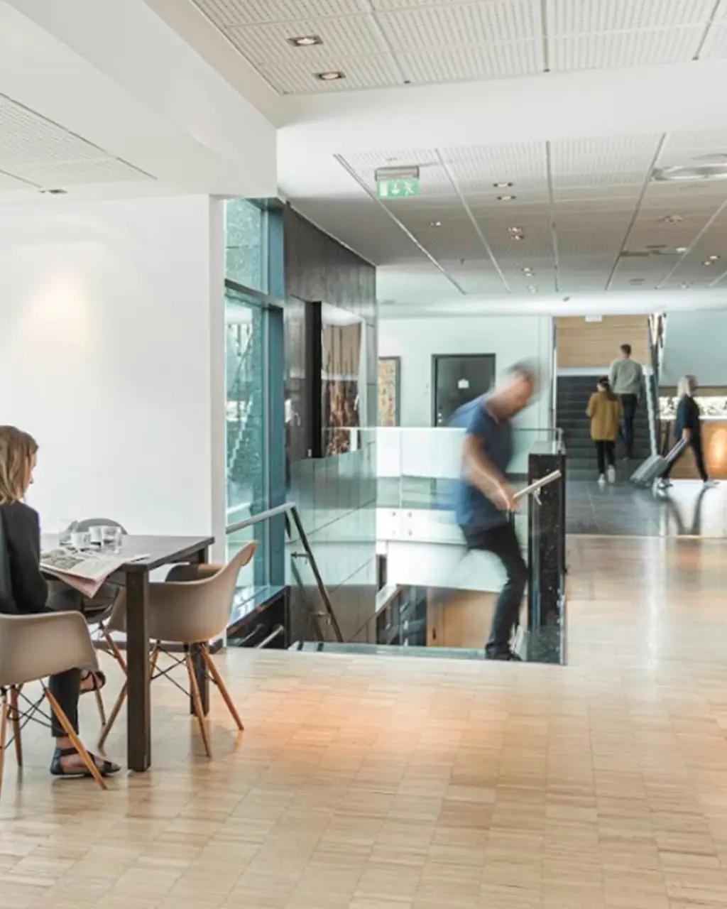Woman reading a newspaper at a table in a bright, modern lobby with blurred people moving, one descending stairs.
