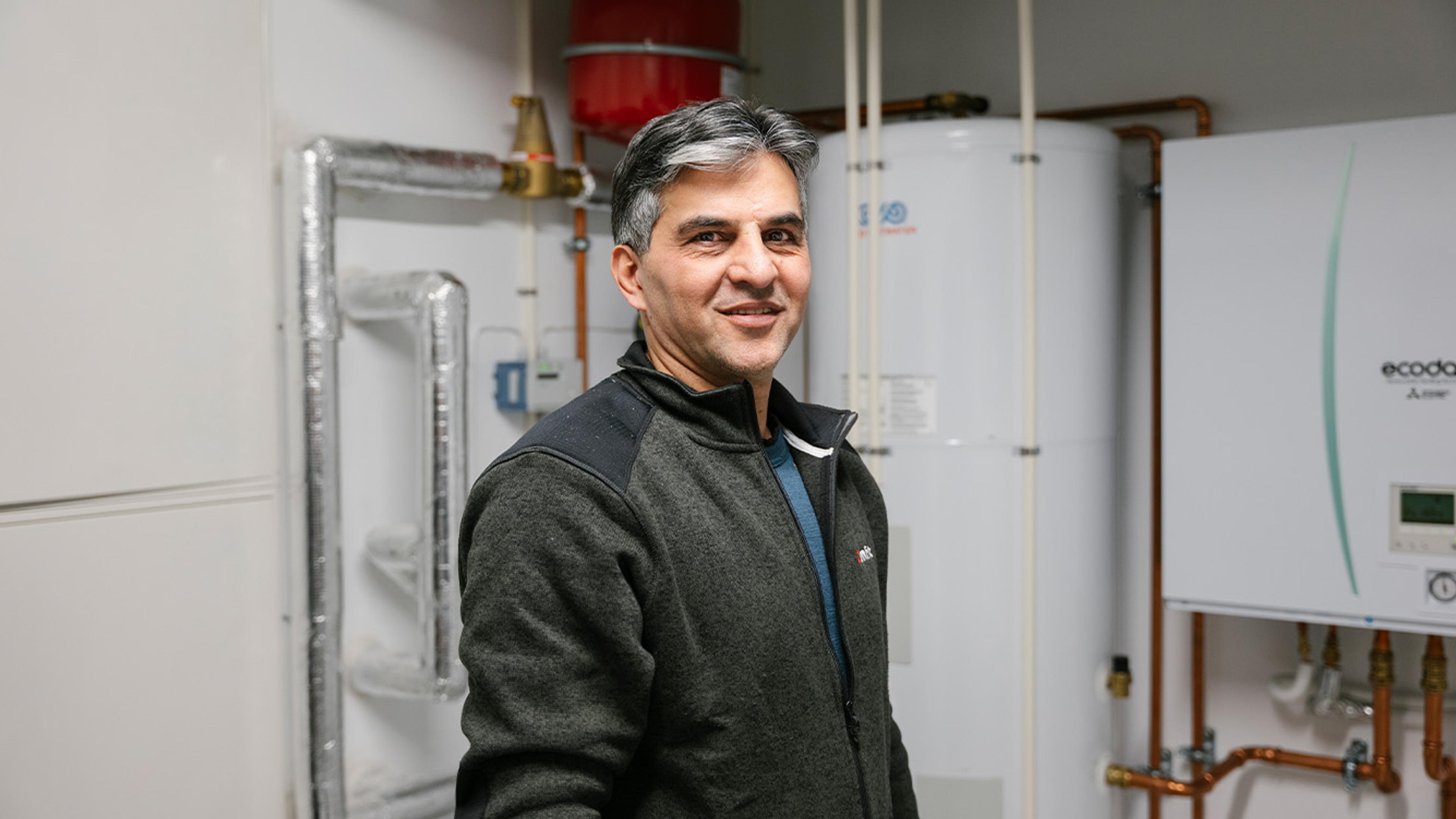 Prosjektleder Vria Naseri foran luft-vann-varmepumpe i teknisk rom