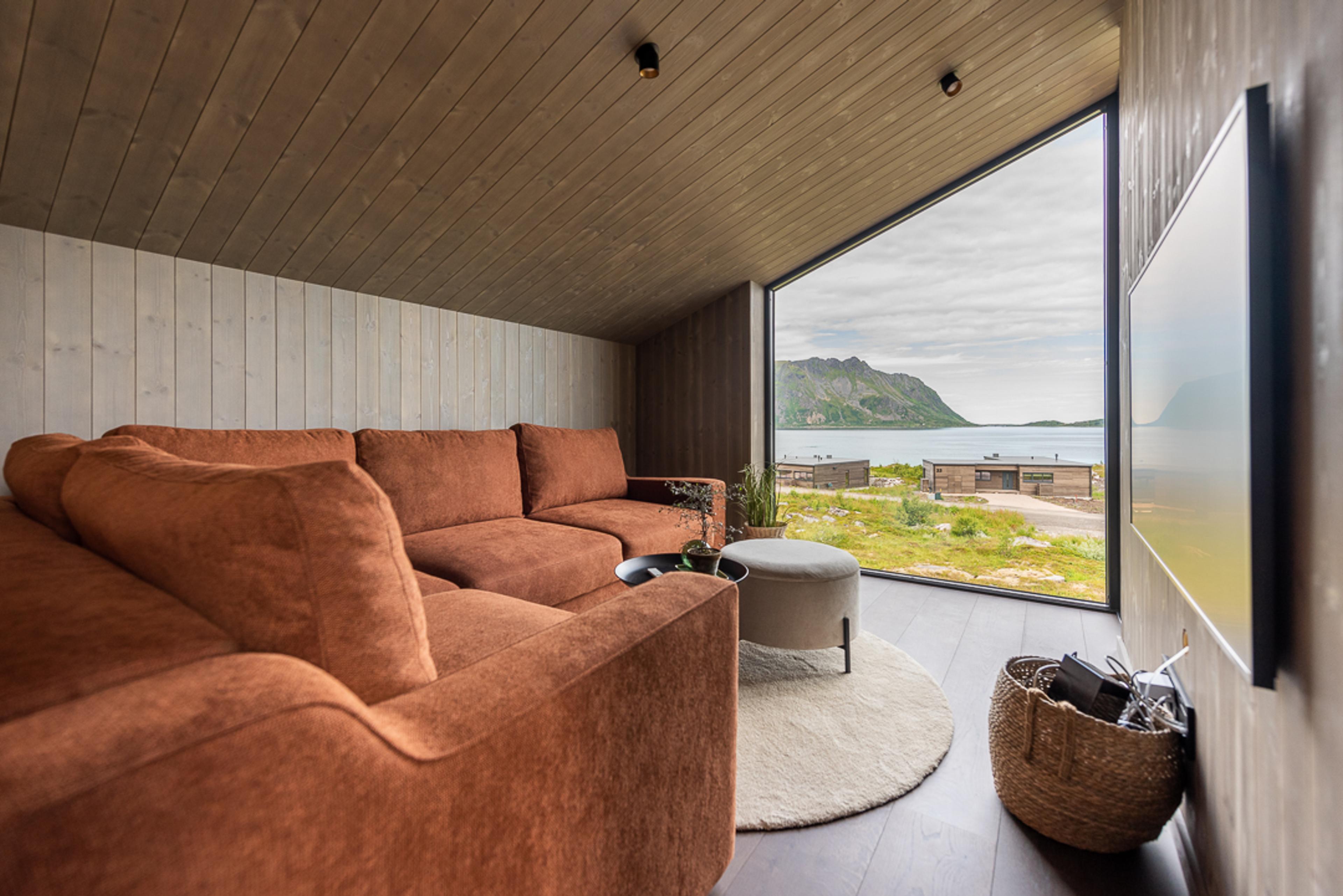 En rustrød sofa i en loftstue i en hytte med vindu og utsikt ut mot havet og fjell