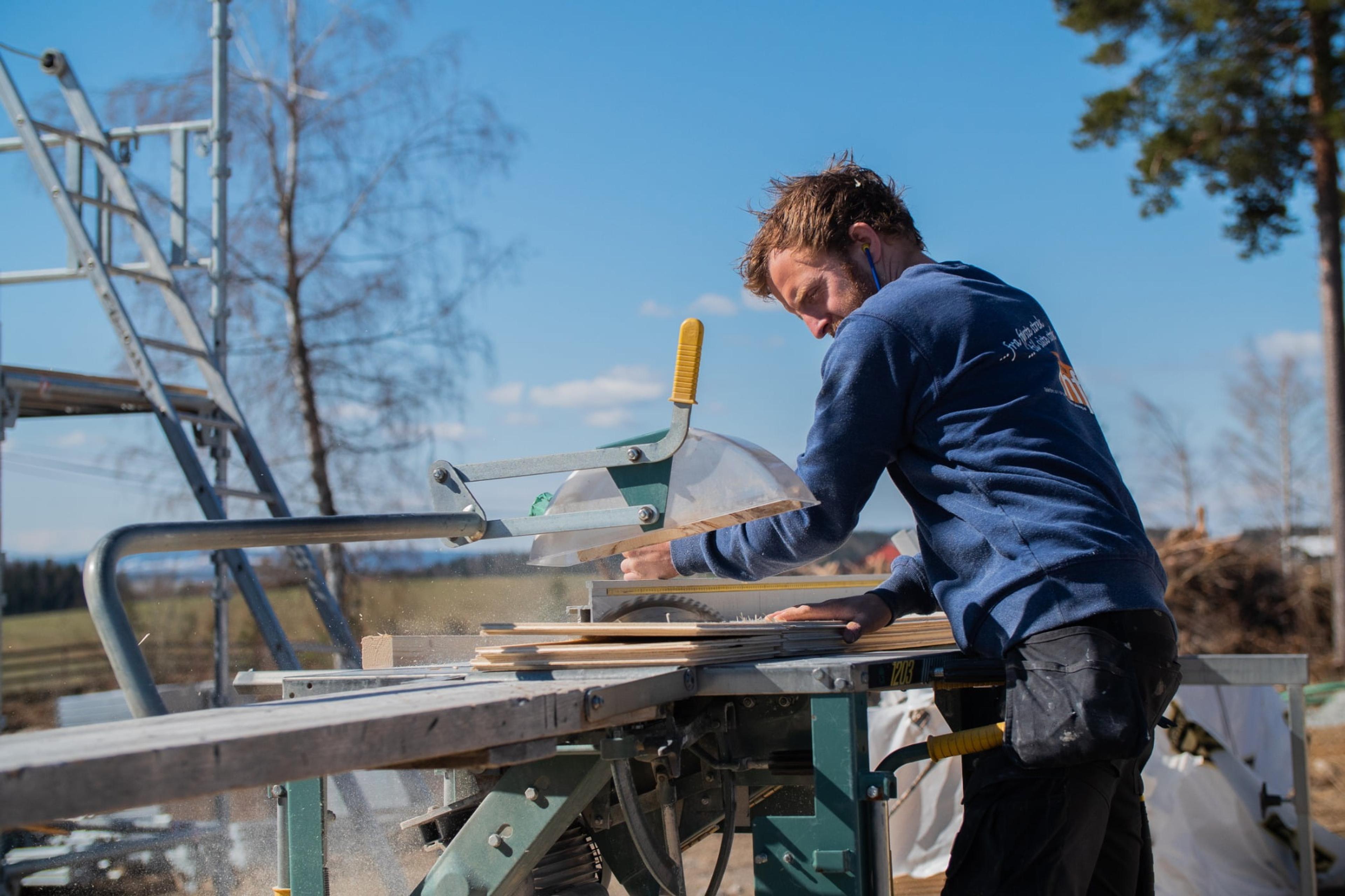 En mann med mørkt hår står ved en sag og kapper plank