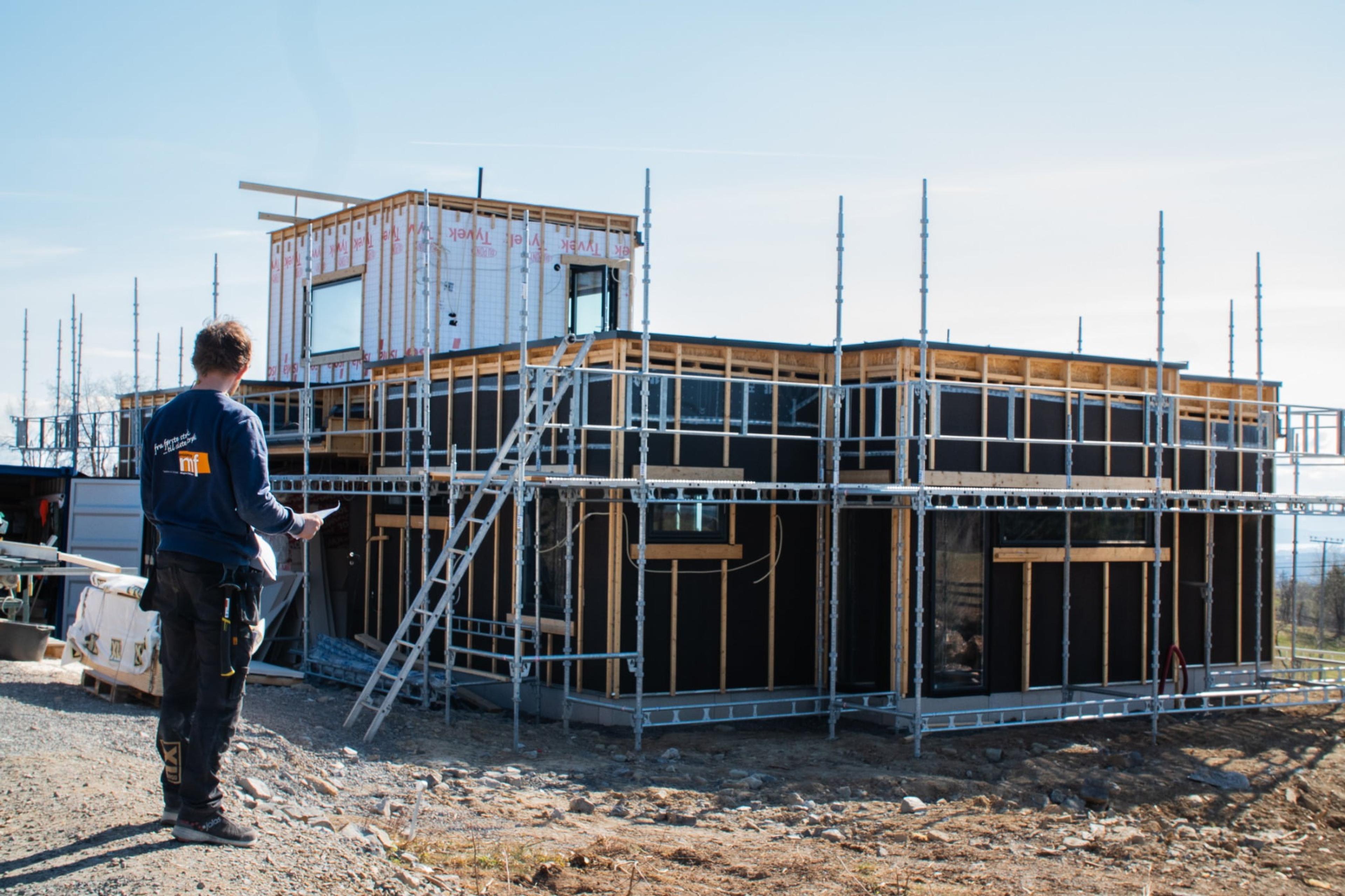 En mann med mørkt hår står ved et hus som holder på å bli bygd
