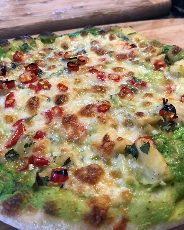 A pizza on a wooden board with a bright green sauce base, melted cheese, sliced red chillies and pineapple