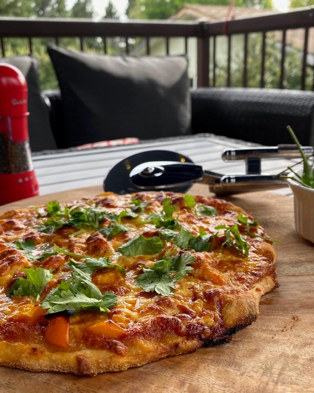 A pizza topped with coriander and orange peppers on a round board on a deck table, a bowl of green chillies beside it