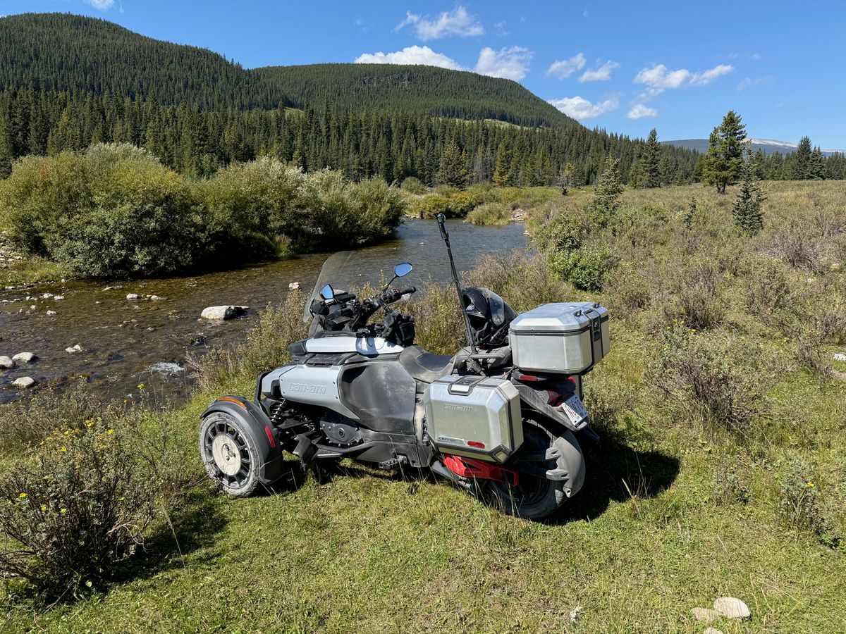 A Can-Am three-wheeler parked in the grass beside a creek in the Rocky Mountains