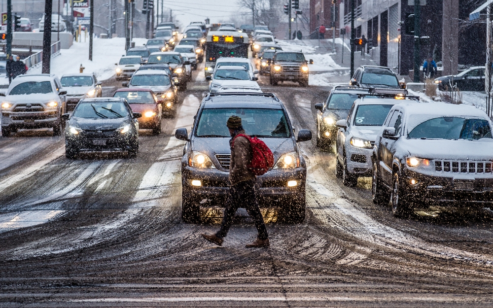 Image A pedestrian with a red backpack crossing a snowy, slushy street during heavy holiday traffic with car headlights reflecting off the wet pavement.