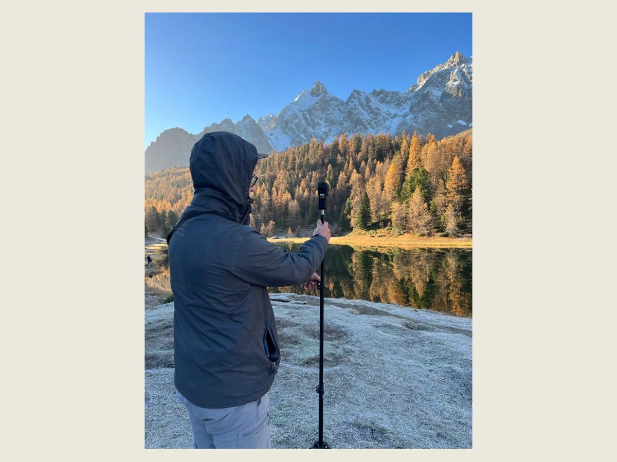 Un technicien de dos, capuche relevée, tient une perche surmontée d'une caméra 360 face au lac Miroir, avec les mélèzes orange et les sommets enneigés