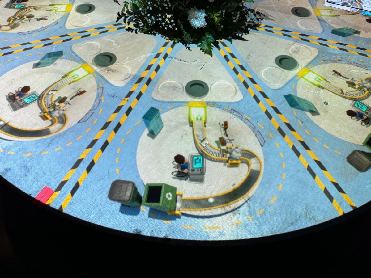 Vue de dessus d’un atelier industriel miniature projeté sur une table ronde : postes de travail, convoyeurs et circulations balisées en jaune et noir